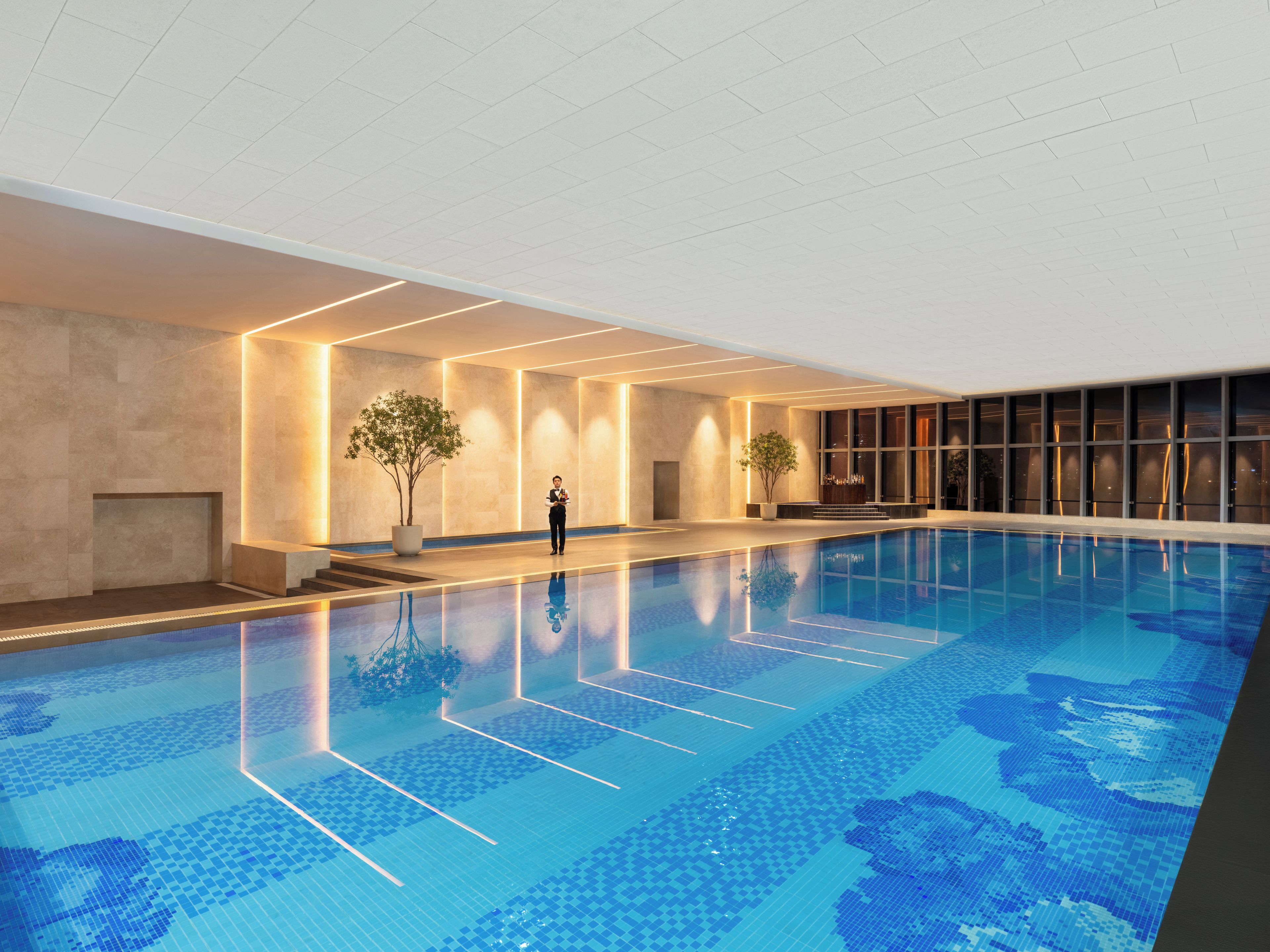 Una piscina interior con suelo de baldosas, una persona de pie junto a la piscina y una zona de bar con un ventanal grande.