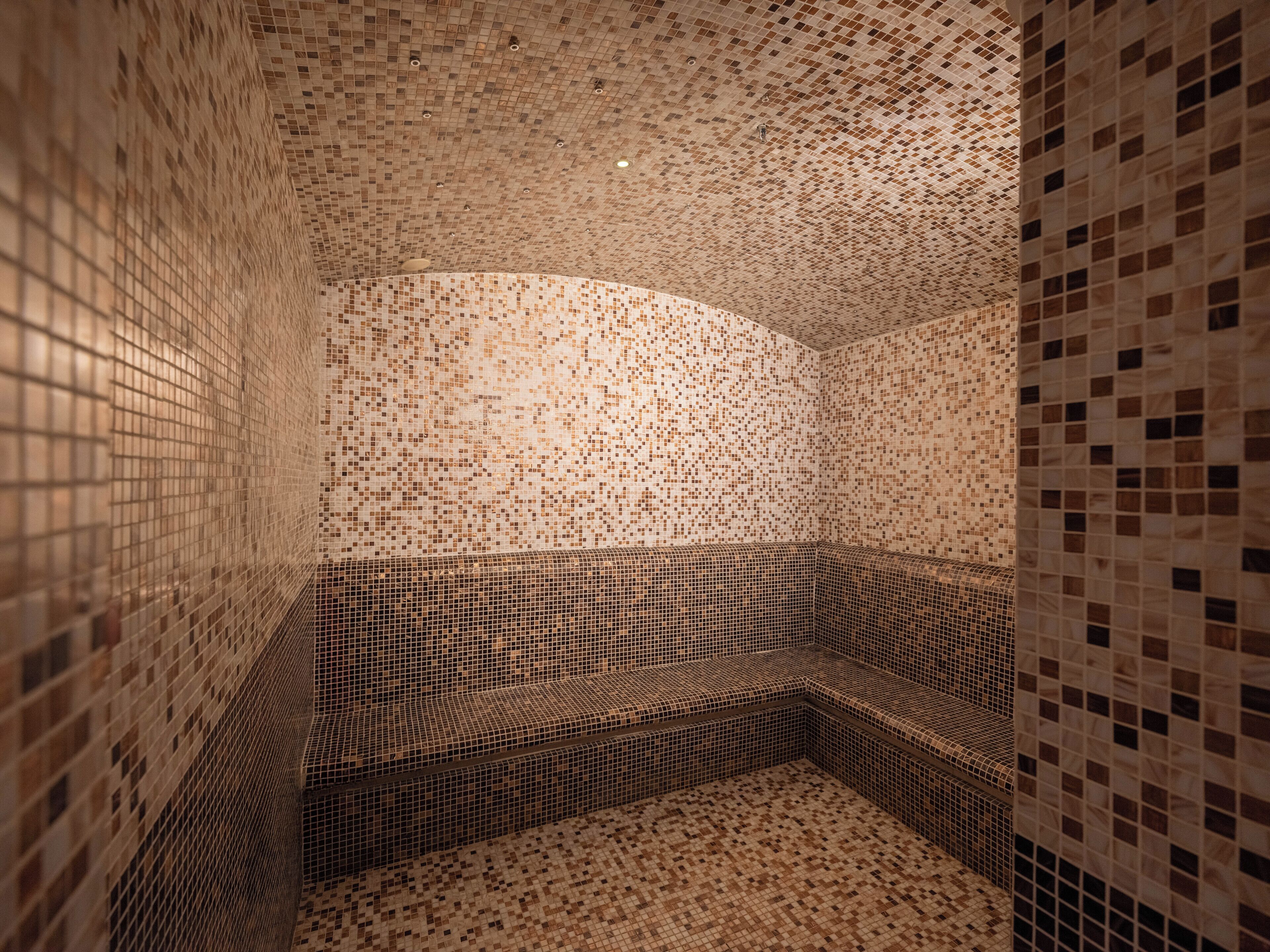 A room with a mosaic-tiled wall and floor, a curved bench, and a ceiling with recessed lighting.