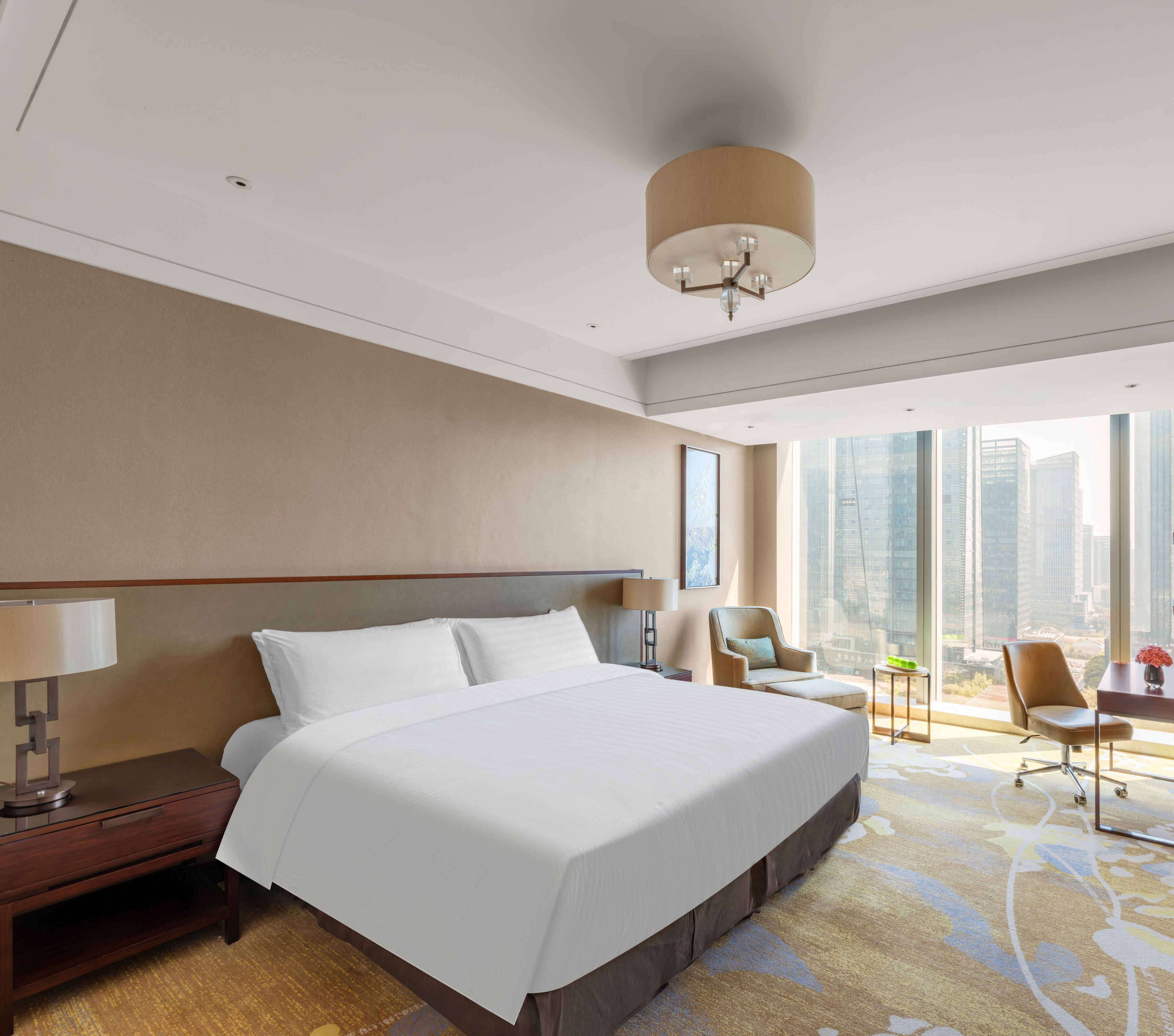 A modern hotel room with a large bed, a desk, and a chair. There is a large window with a city view.