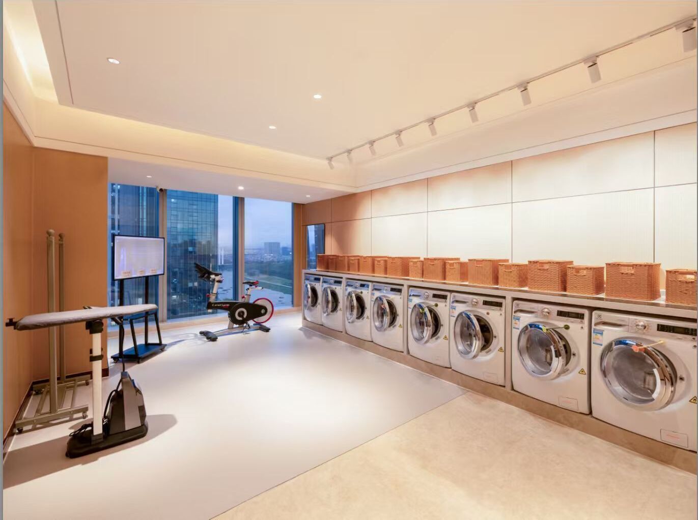 A modern laundry facility with multiple washing machines, a treadmill, and a large window offering a city view.