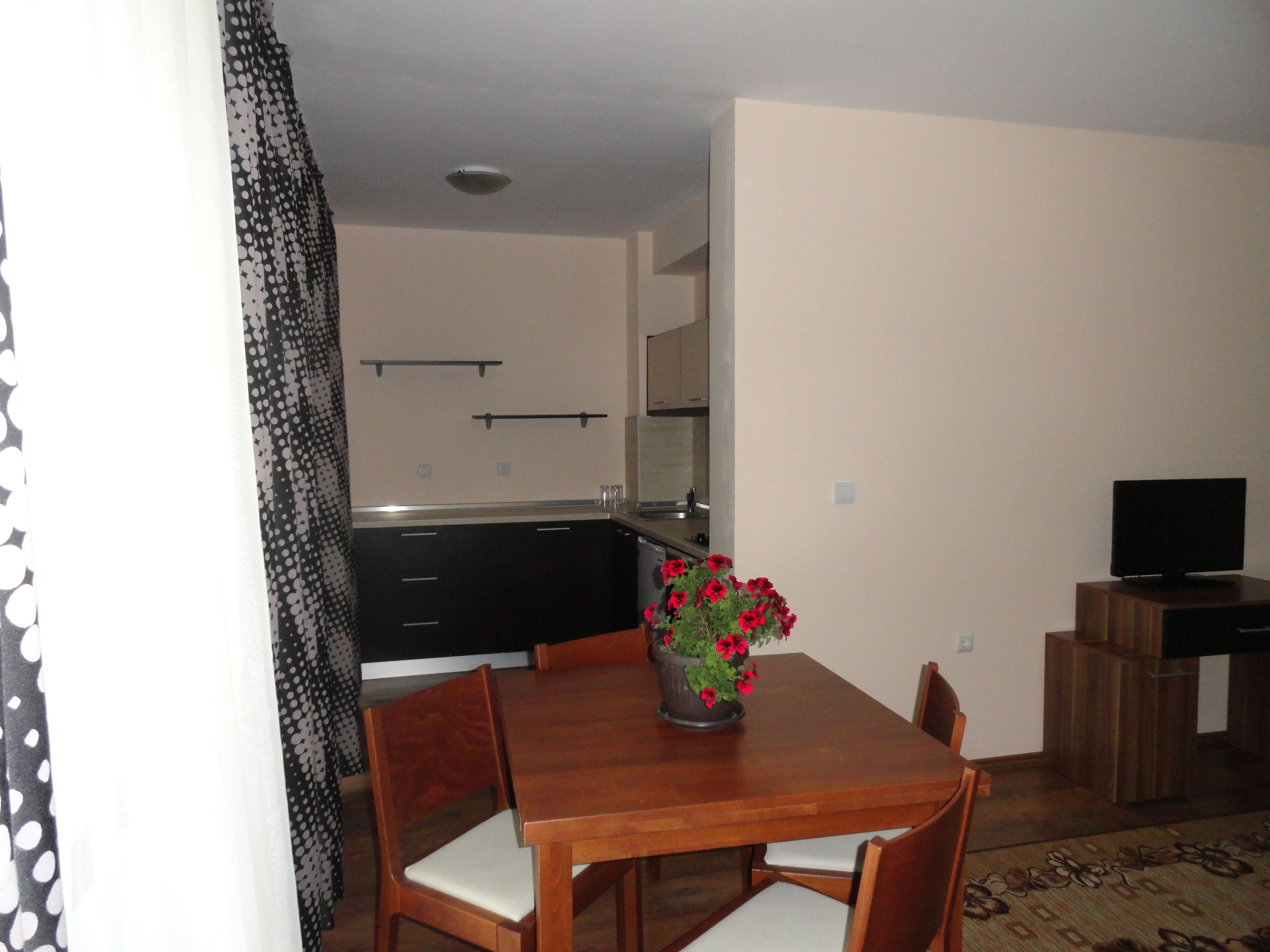 A dining area with a wooden table, four chairs, a vase with red flowers, a television on a wooden stand, and a kitchen in the background.