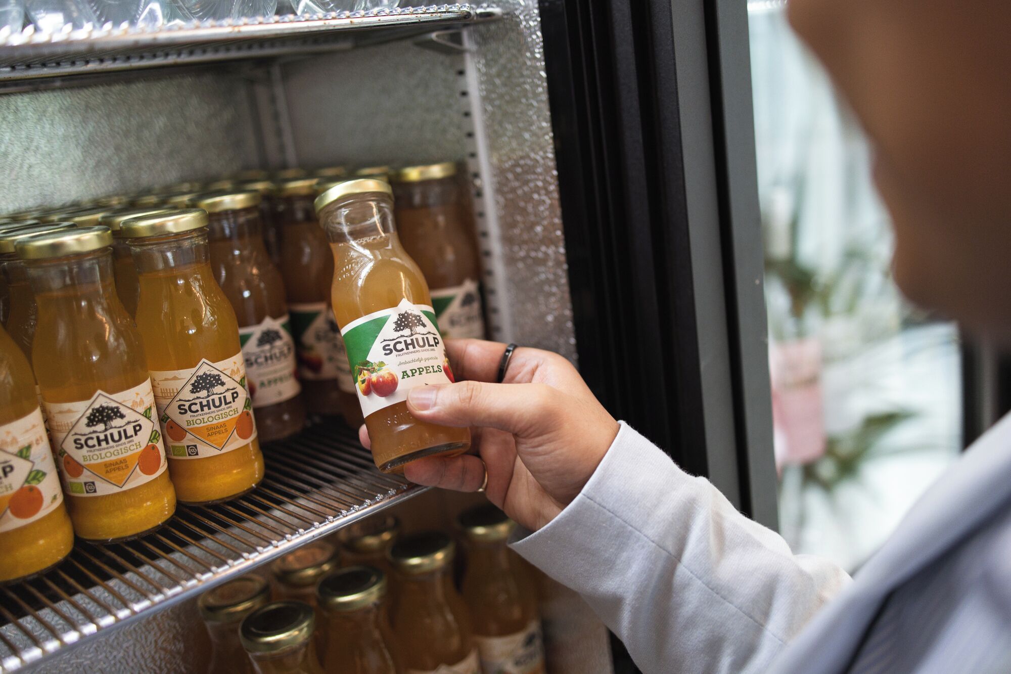 A hand selecting a bottle of SCHULP apple juice from a refrigerated display.