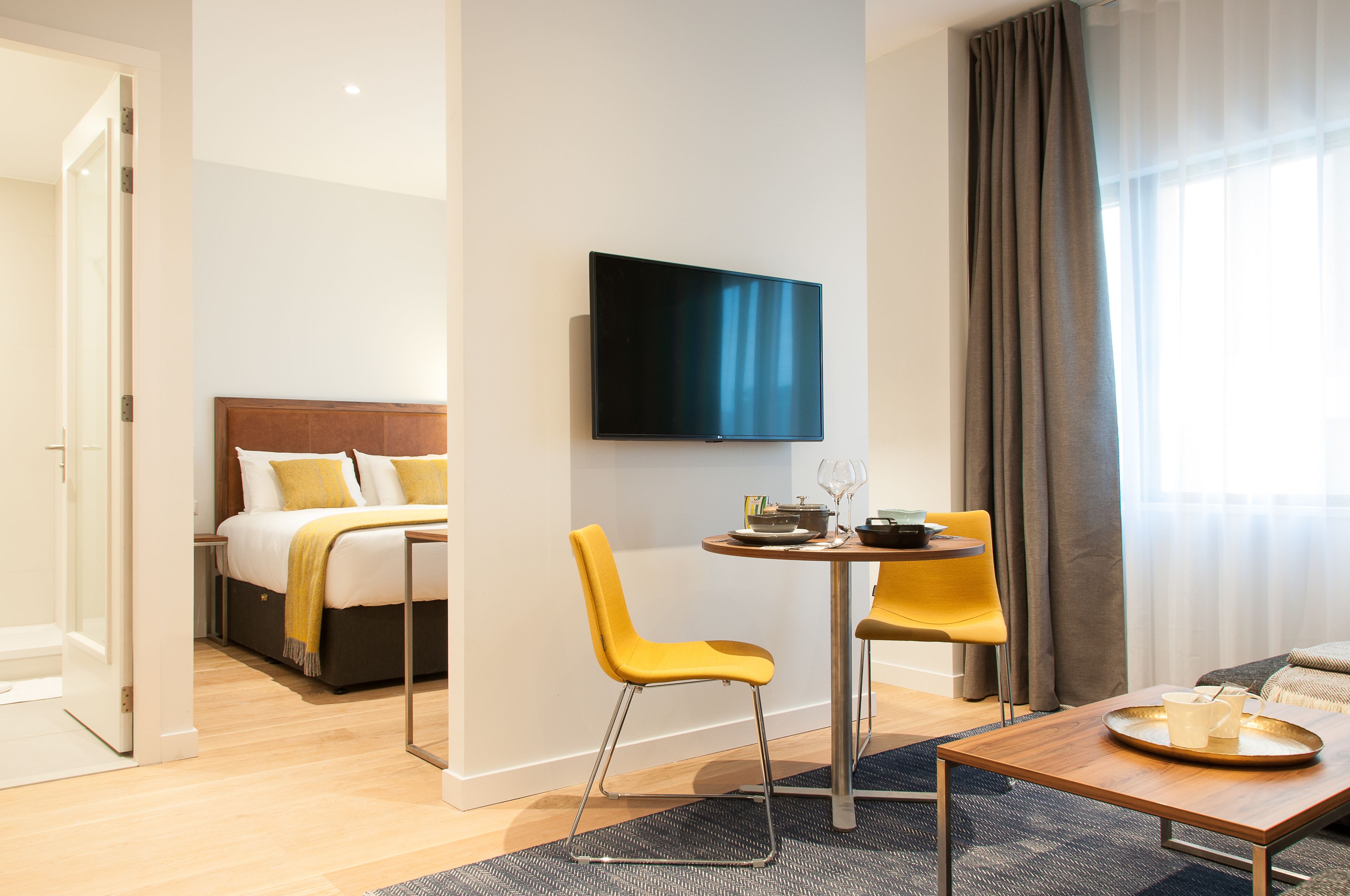 Una camera d'albergo con un letto, una televisione a parete, un tavolo da pranzo con sedie e una finestra con tende.