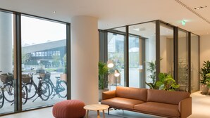 En modern inredning med en lädersoffa, en rund pall och ett litet bord. Stora fönster ger utsikt över cyklar och en byggnad utanför.