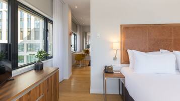 Habitación de hotel con un escritorio de madera, una cama con sábanas blancas, una mesita de noche con una lámpara y vistas a la ciudad a través de grandes ventanales.