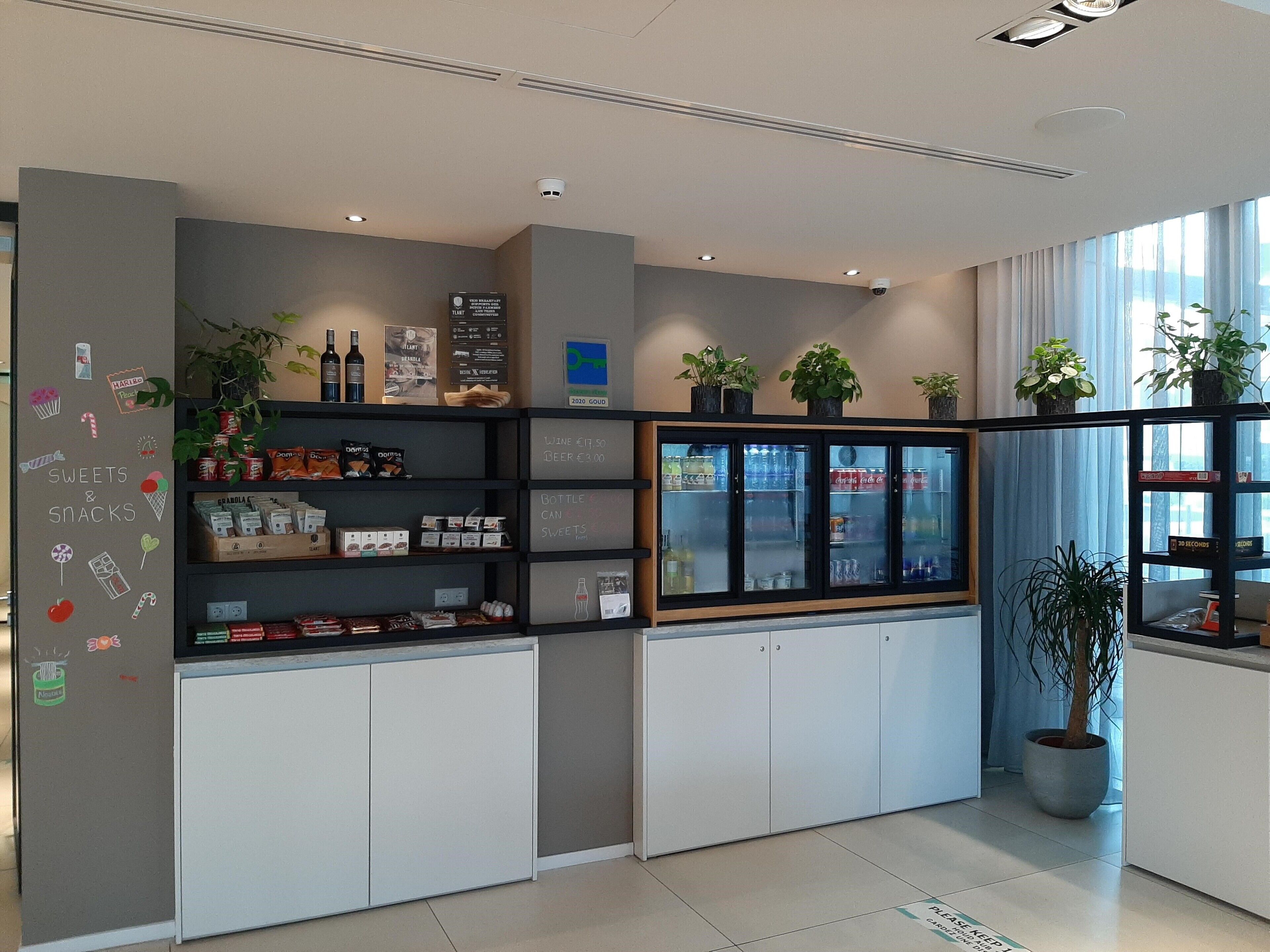 A modern interior with shelves stocked with snacks and beverages, a refrigerated display case, and potted plants.