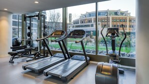 Ett gym med löpband, styrketräningsmaskiner och en roddmaskin, med stora fönster som ger utsikt över en byggnad och grönområden utanför.