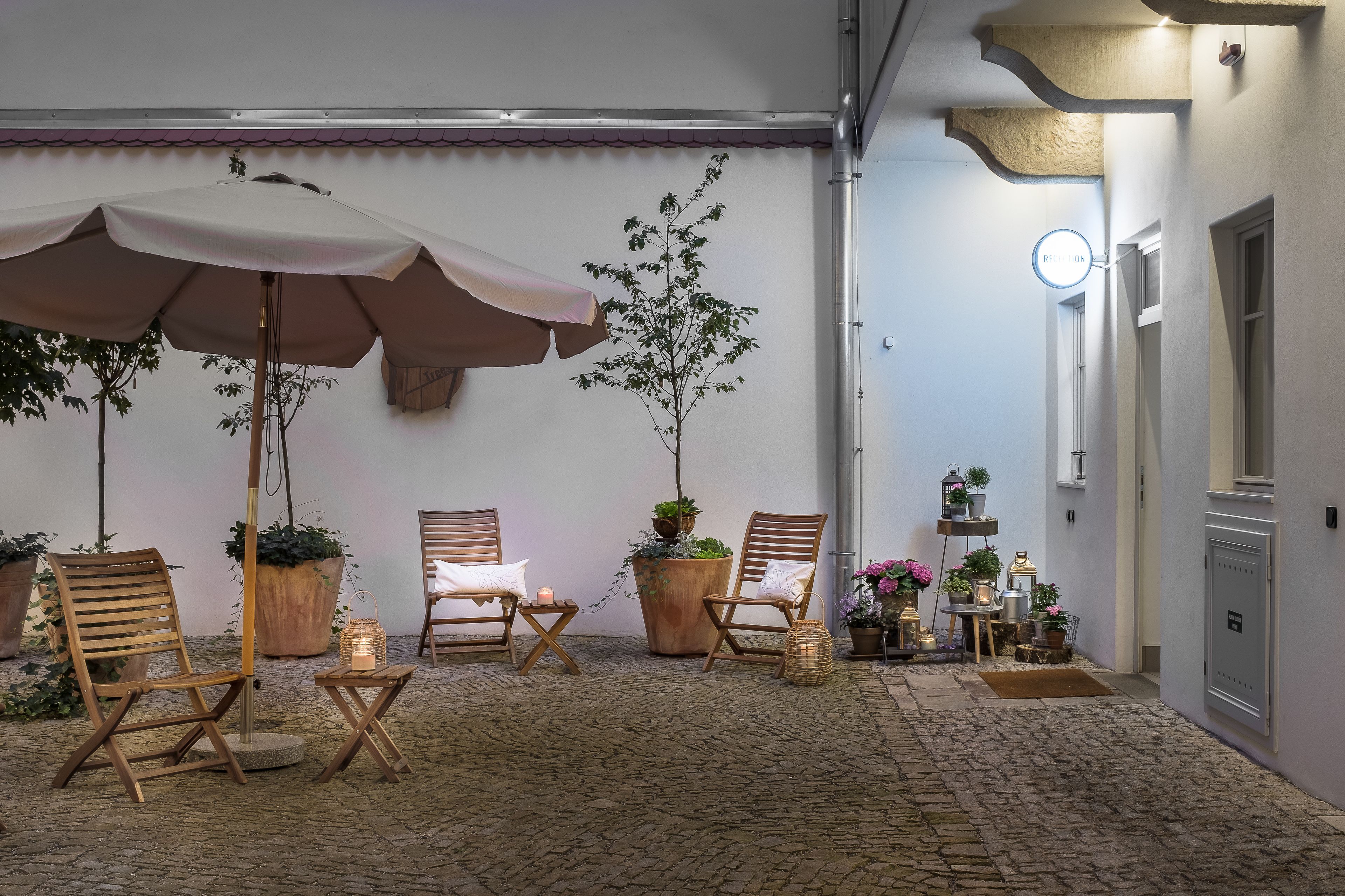 Een gezellig buitenzitgedeelte met houten stoelen en een grote parasol, omgeven door potplanten en een stenen vloer.