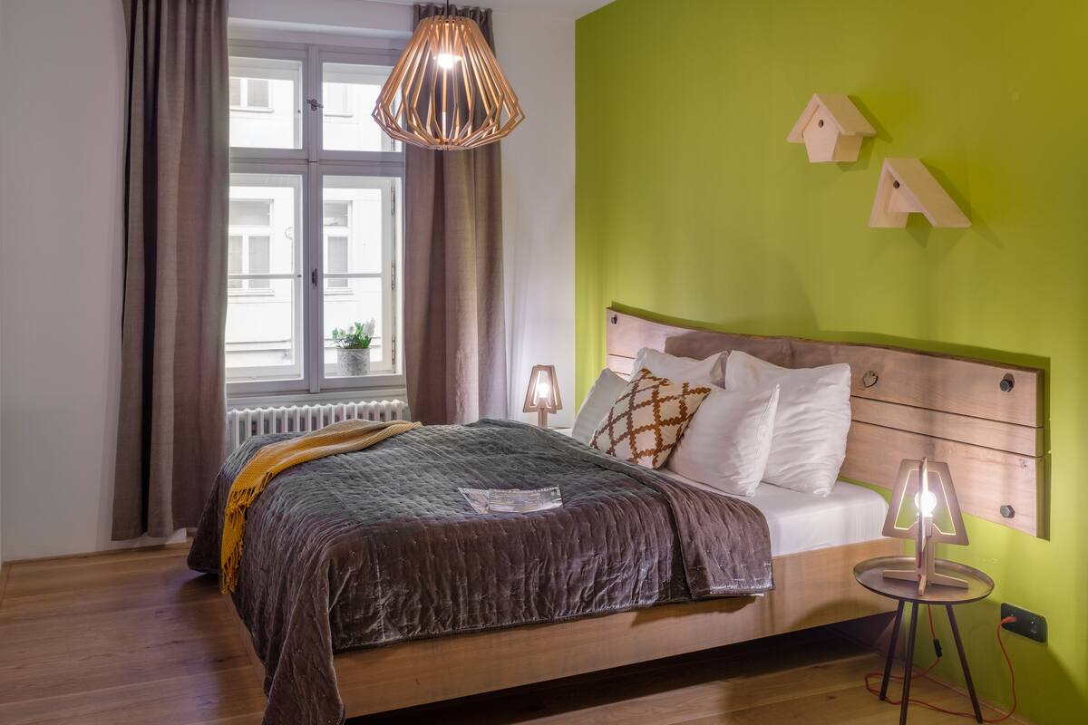 Um quarto com cama de madeira, paredes verdes, uma janela com cortinas, abajur na mesa de cabeceira e uma pequena mesa com luminária.
