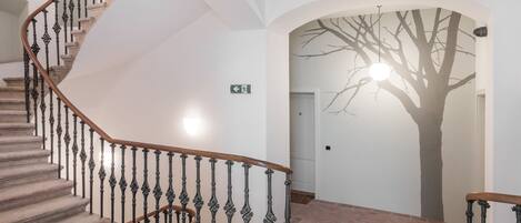 Un escalier en colimaçon orné de rampes noires et d’une main courante en bois, menant à un palier avec un mur blanc et une fresque représentant un arbre.