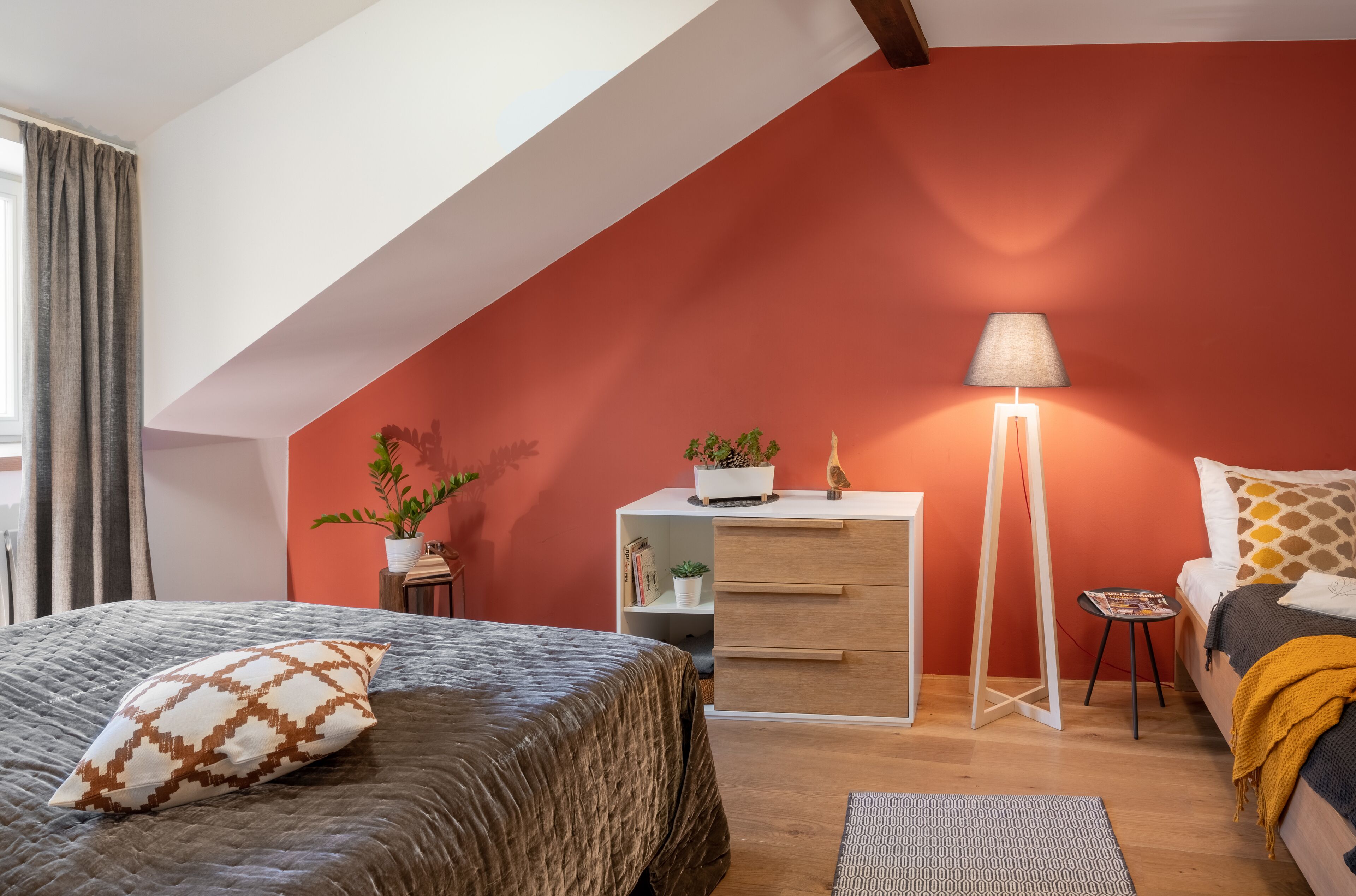 Una camera da letto con soffitto spiovente, un letto con cuscini decorati, un comodino con una lampada, una cassettiera e un piccolo tavolino con una pianta.