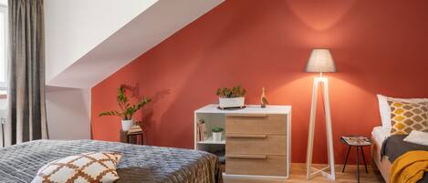 Una camera da letto con soffitto spiovente, un letto con cuscini decorati, un comodino con una lampada, una cassettiera e un piccolo tavolino con una pianta.