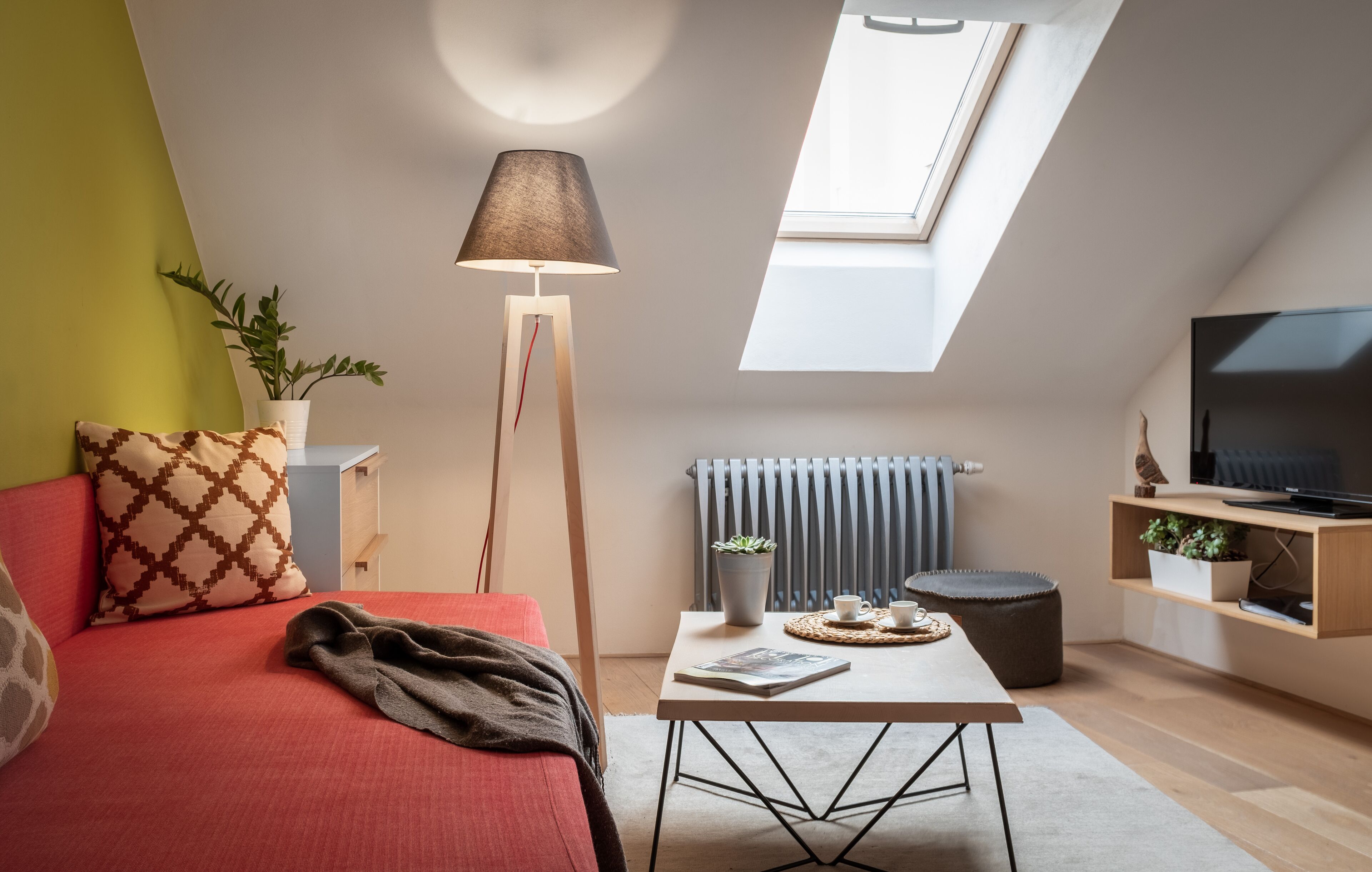 A cozy loft bedroom with a red sofa, a floor lamp, a small table with a magazine, and a TV on a wooden shelf.