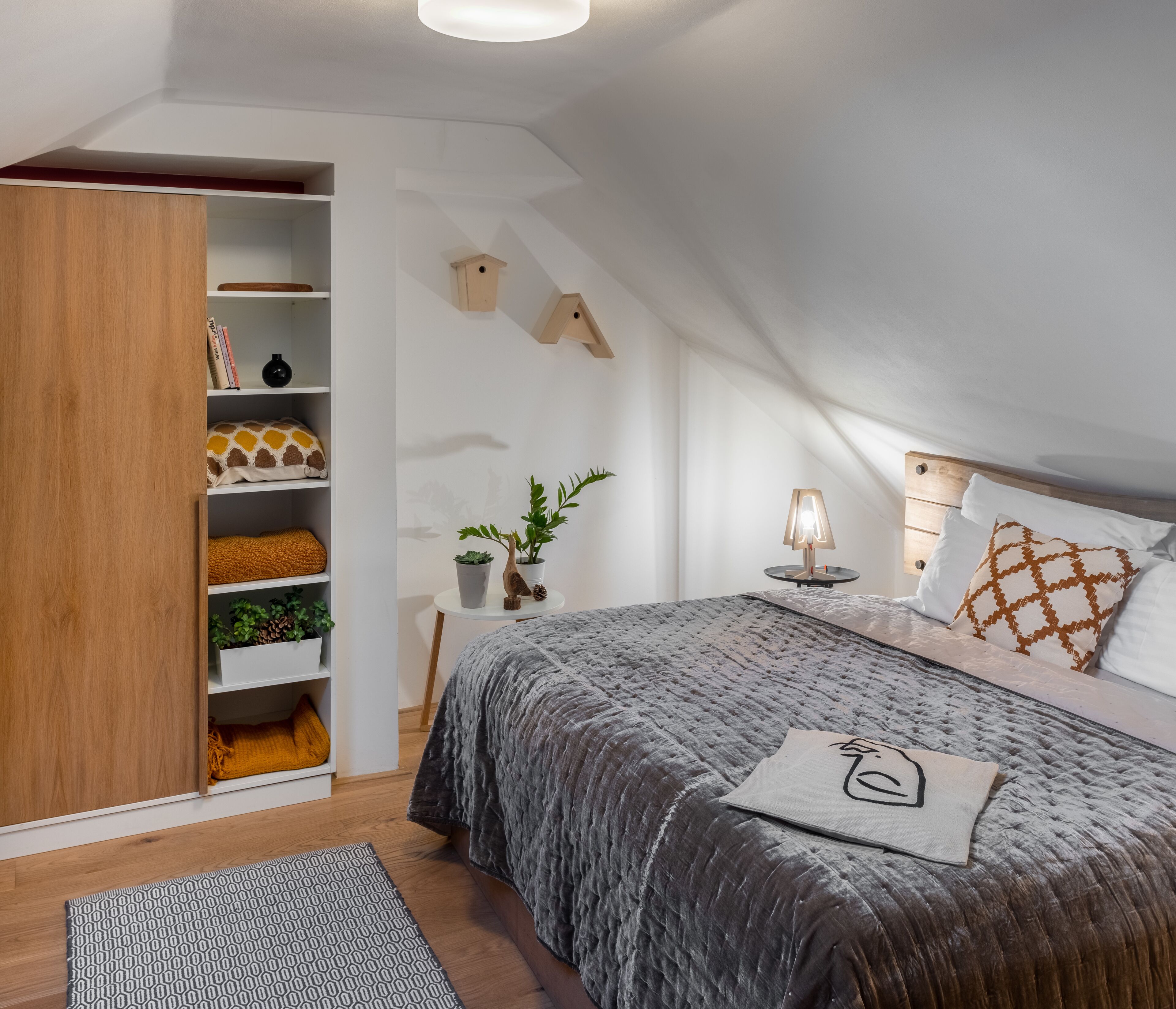 A bedroom with a loft-style ceiling, a bed with a grey quilt, a bedside table with a lamp, a wooden wardrobe, and a shelving unit with books and decorative items.