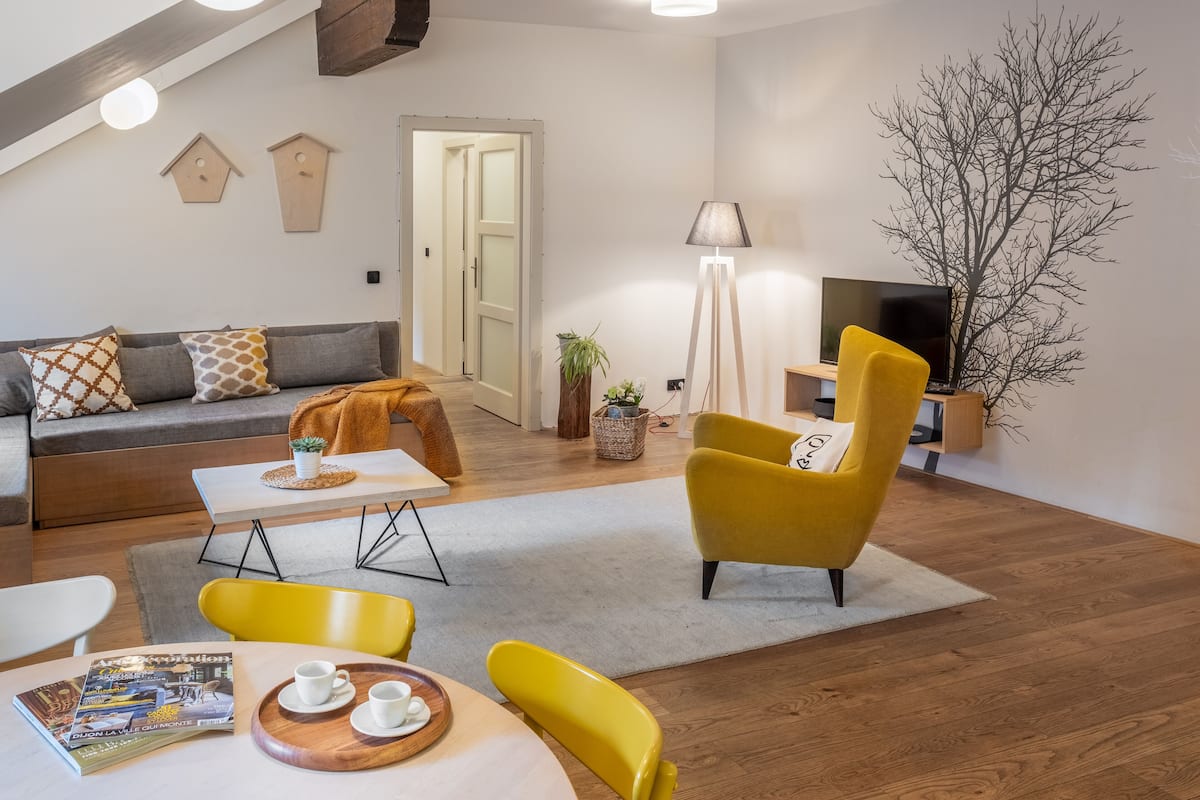 Uma sala de estar moderna com sofá, cadeiras amarelas, uma mesa de jantar com revista e xícaras, luminária, televisão e planta.