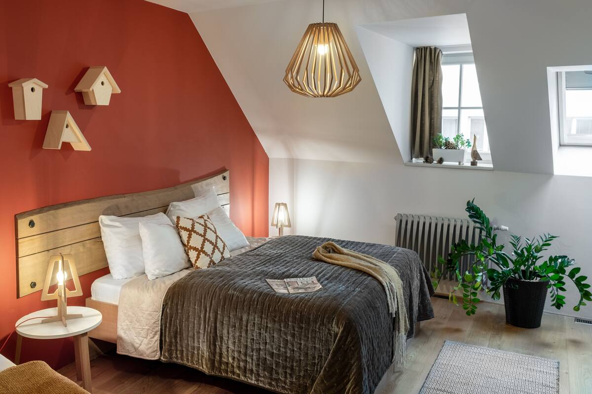Um quarto com teto inclinado, uma cama com cabeceira de madeira, uma mesa de cabeceira com abajur, um radiador e uma janela com vista para o exterior.