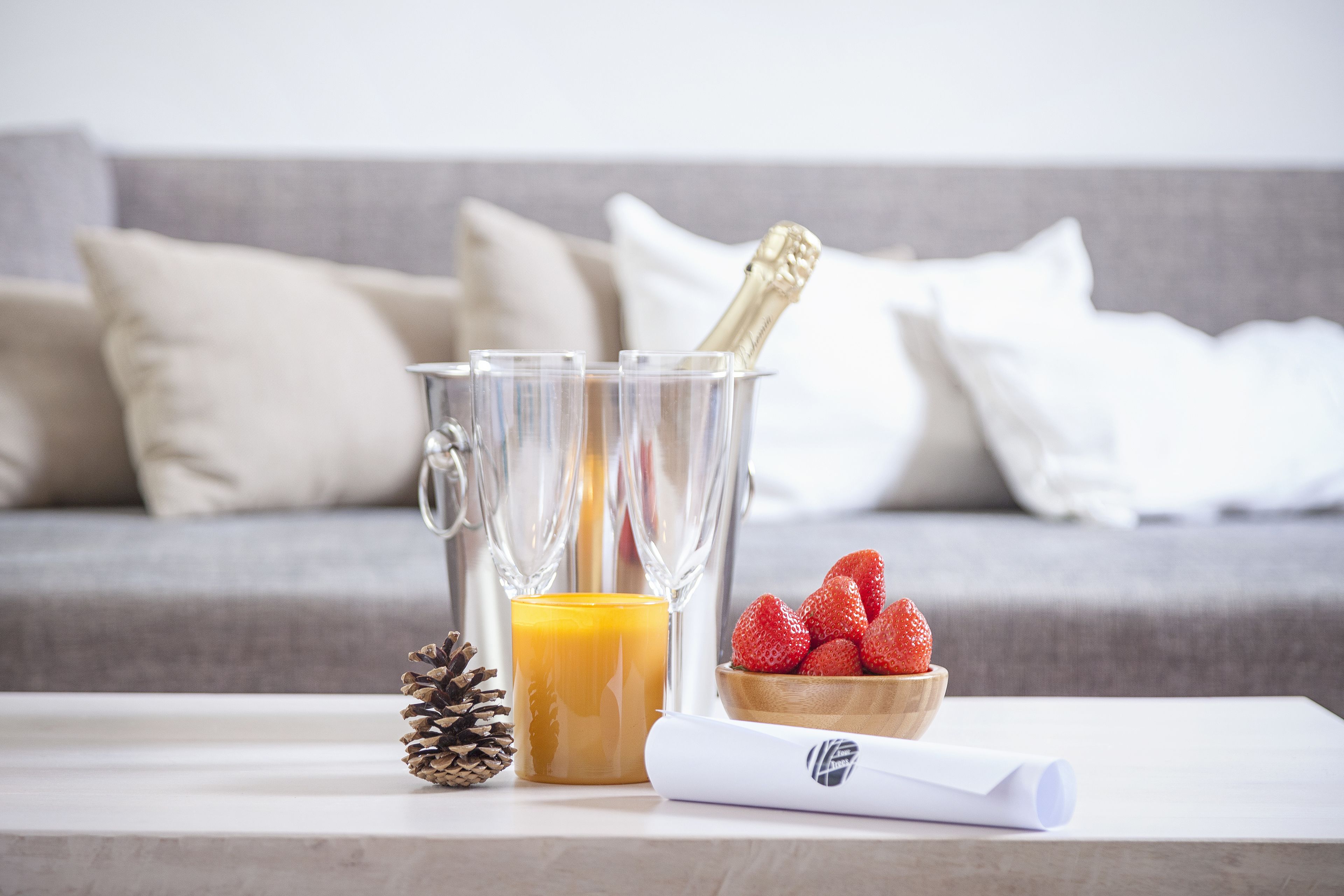 A clear glass decanter with a bottle of champagne, a glass of orange juice, a pine cone, a bowl of strawberries, and a rolled-up piece of paper on a white table in front of a grey sofa with white cushions.