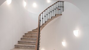 Uma escadaria em espiral com degraus de pedra e guarda-corpo de madeira, conduzindo a um patamar com parede branca e uma pequena arcada.