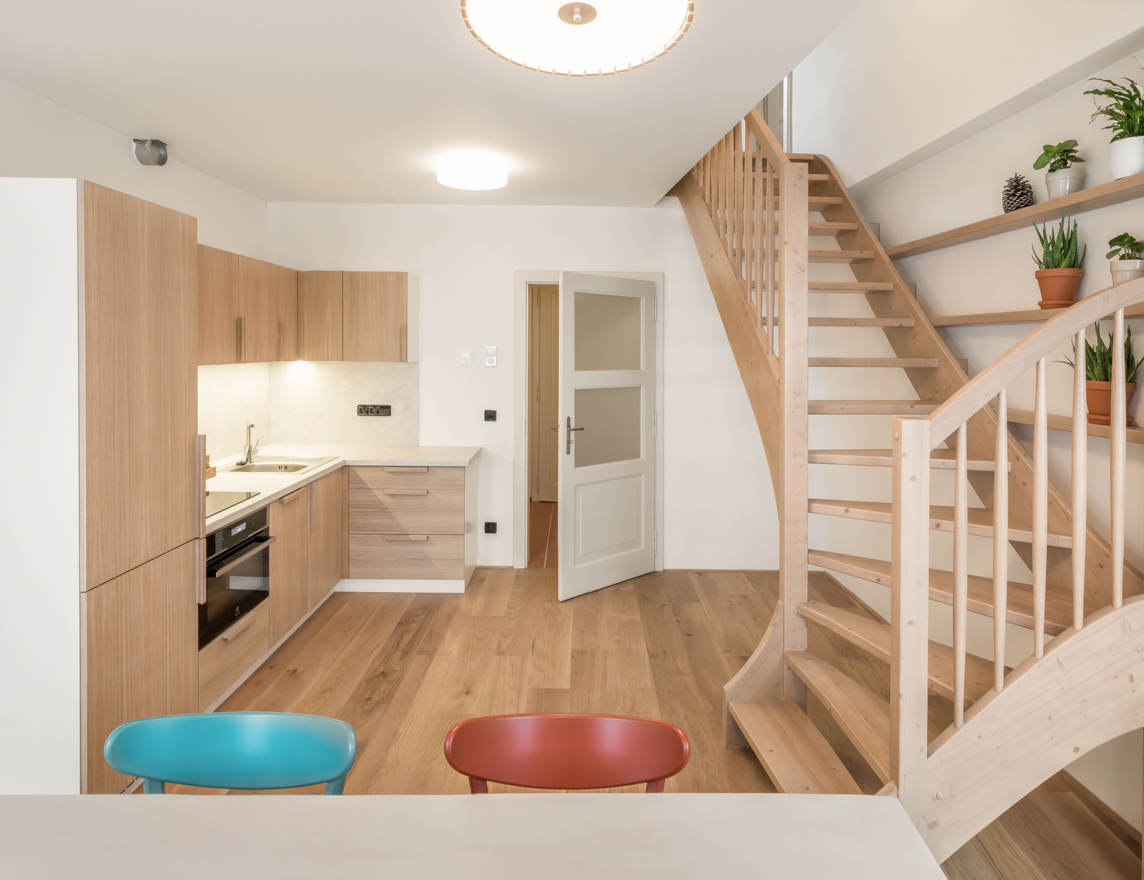 Une cuisine moderne avec des armoires en bois, un four encastré et un évier. Un escalier avec des rampes en bois mène à un palier agrémenté de plantes en pot. On aperçoit deux chaises colorées au premier plan.