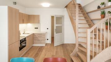 A modern kitchen with wooden cabinets, a built-in oven, and a sink. There is a staircase with wooden railings leading to a landing with potted plants. Two colorful chairs are visible in the foreground.