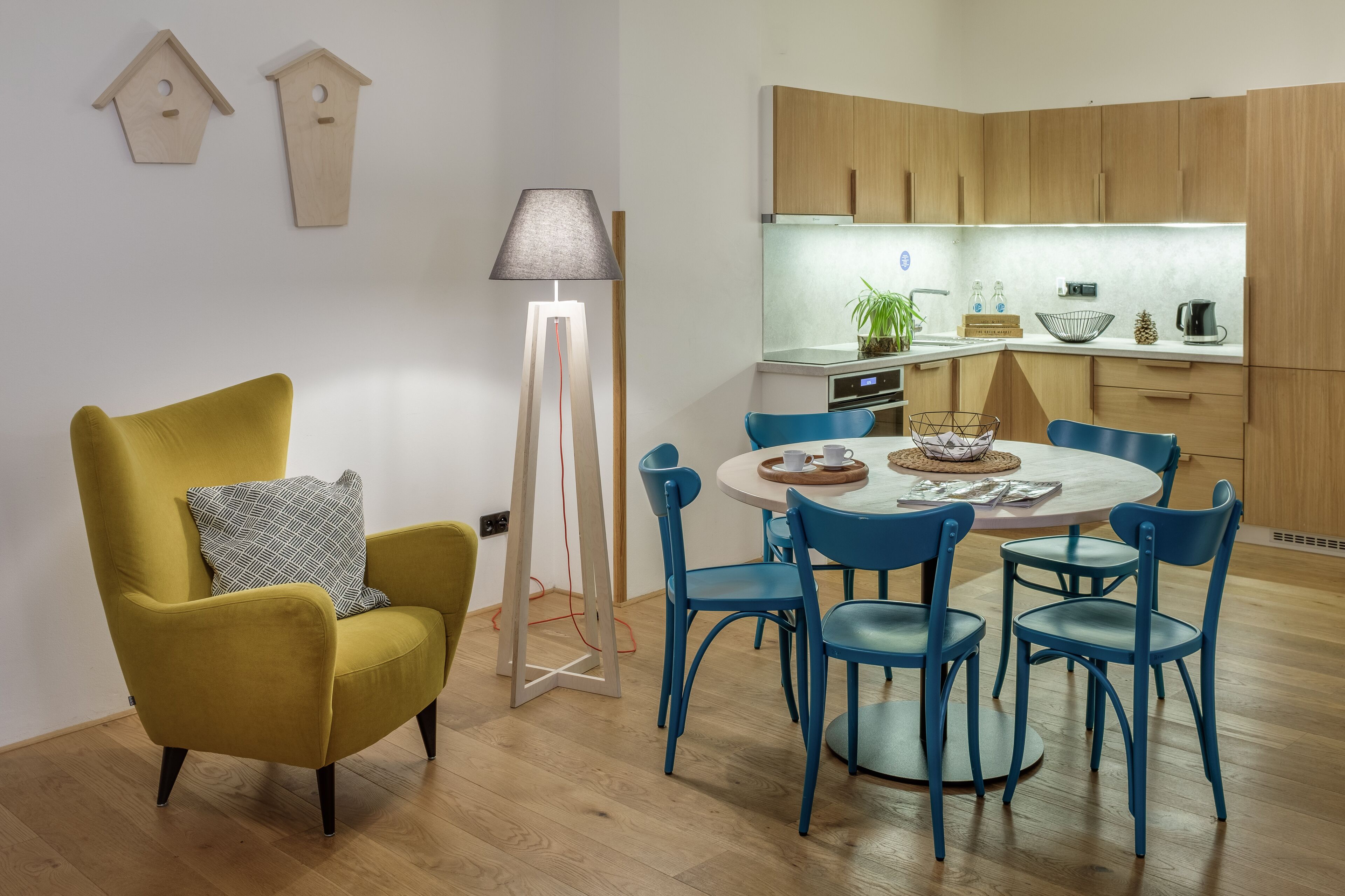 A modern kitchen with wooden cabinets, a round dining table with blue chairs, and a yellow armchair with a patterned pillow.