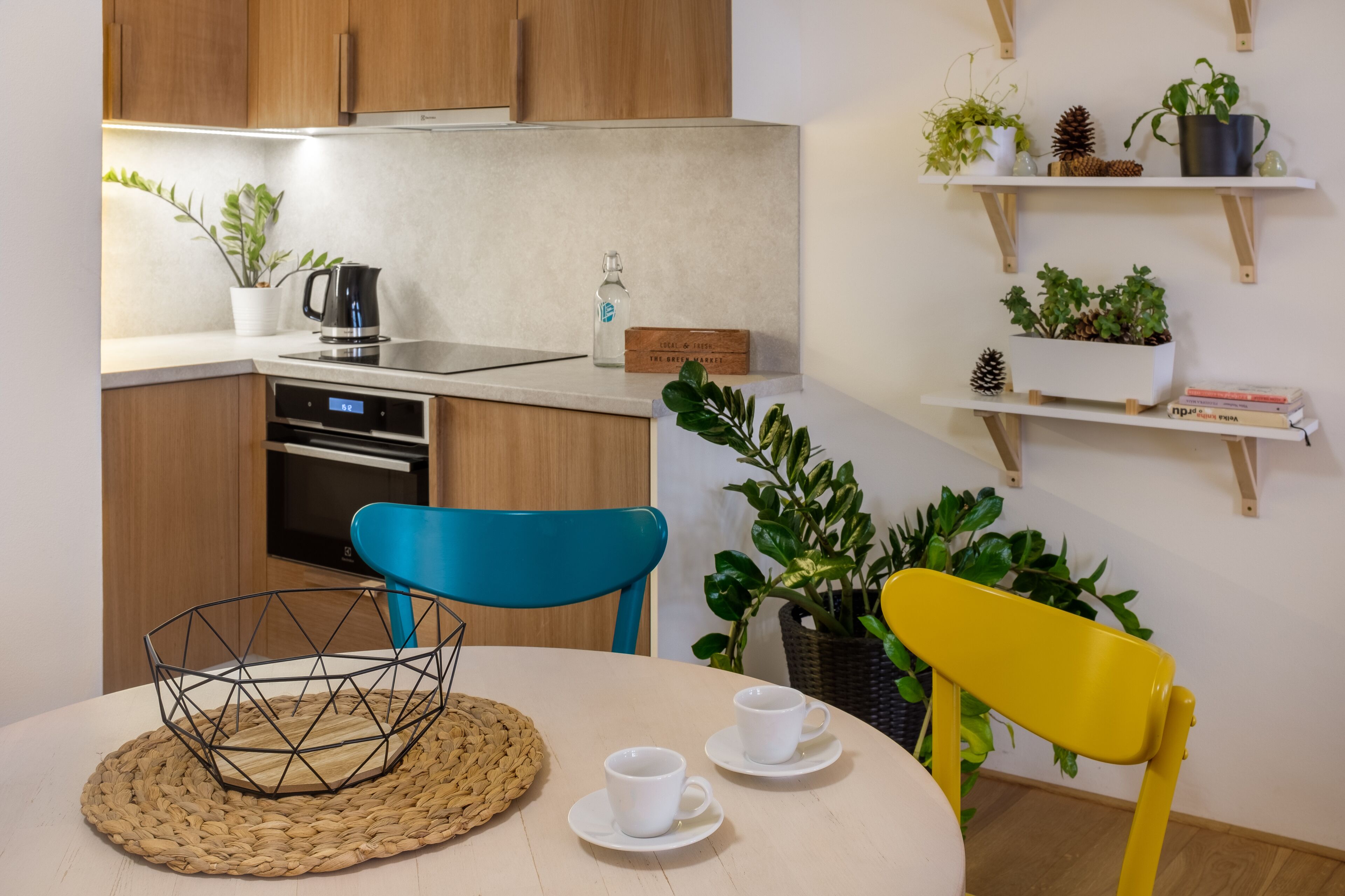 A modern kitchen with wooden cabinets, a built-in oven, a coffee maker, and a dining area with a round table, two chairs, and a woven placemat.