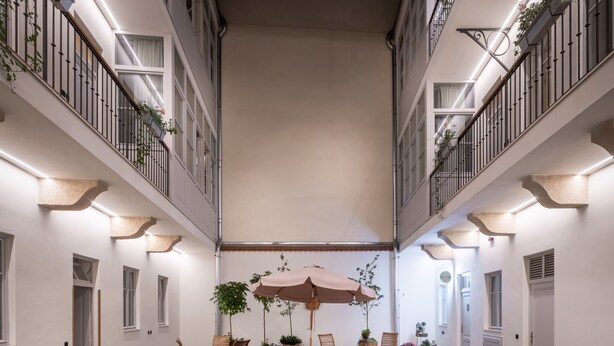 Um pátio espaçoso com área de estar central, com cadeiras e mesas de madeira, rodeado por plantas em vasos e um grande guarda-sol.
