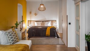Quarto moderno com cabeceira de madeira, cama grande com cobertura escura, mesa de cabeceira com abajur e luminária pendente.