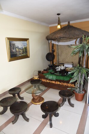 A room with a bed, a painting, a thatched roof, a potted plant, and a small table with stools.