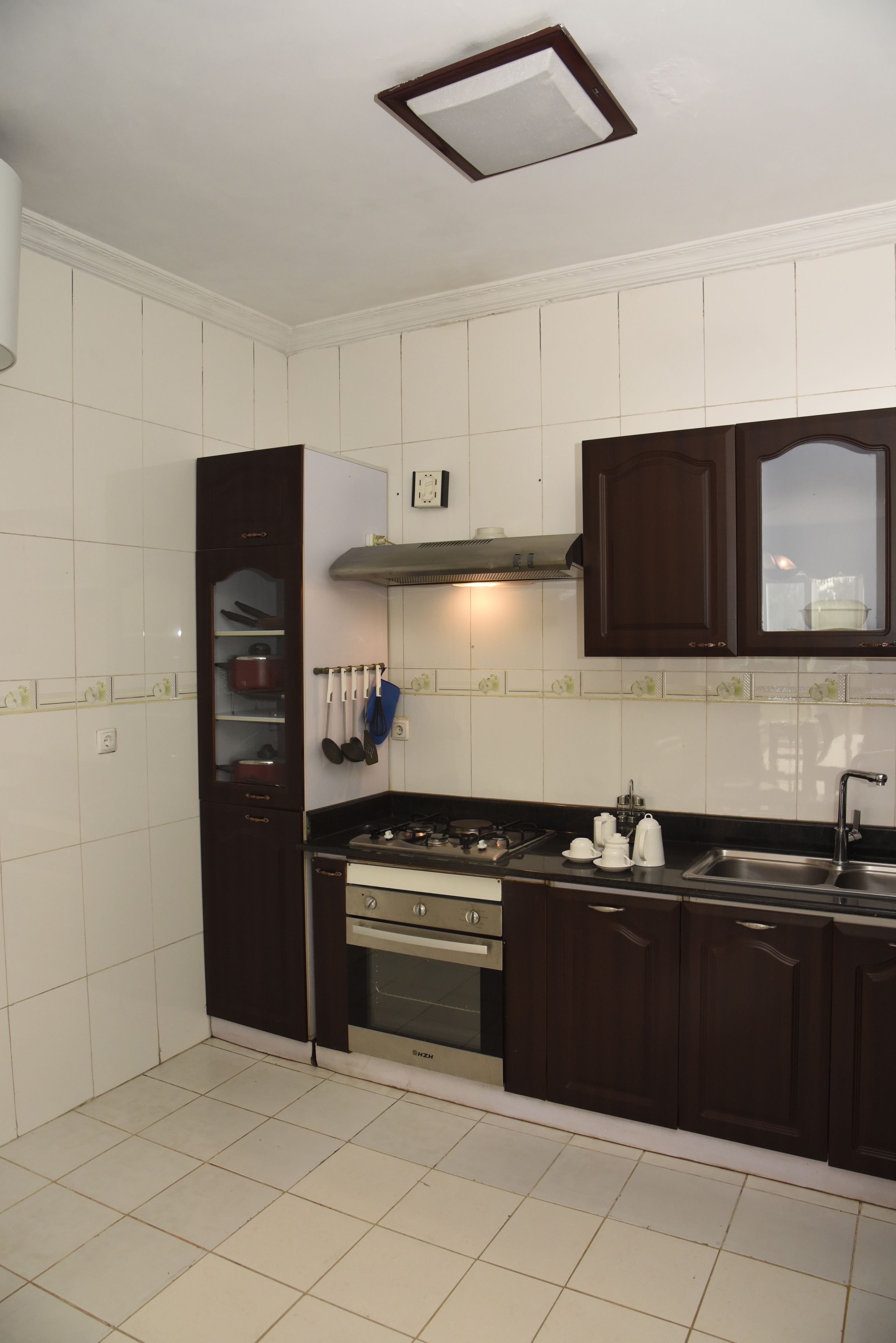 A kitchen with white tiled walls, dark wooden cabinets, a gas stove, and a stainless steel sink.