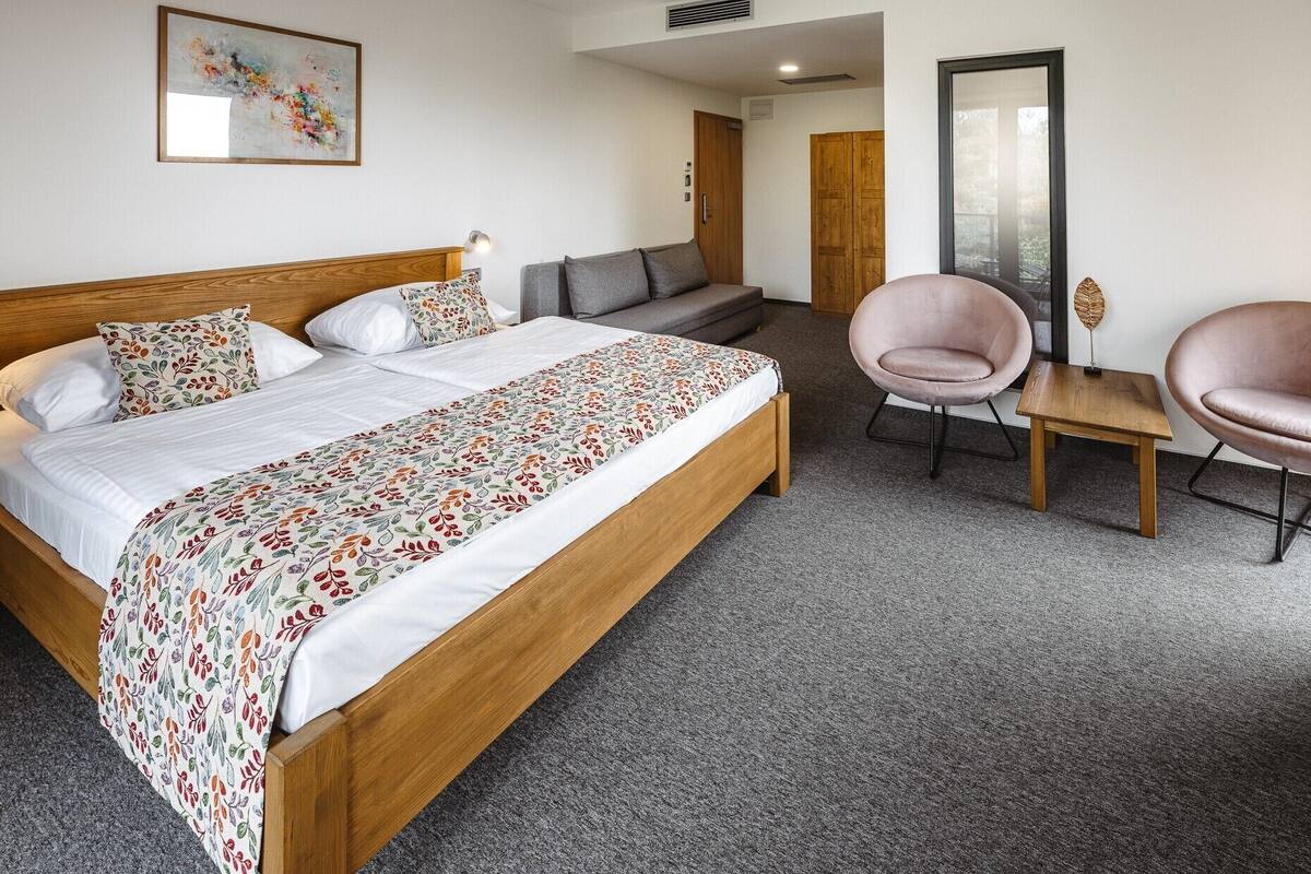 A hotel room with a wooden bed, grey sofa, two pink chairs, a small wooden table, and a large mirror.