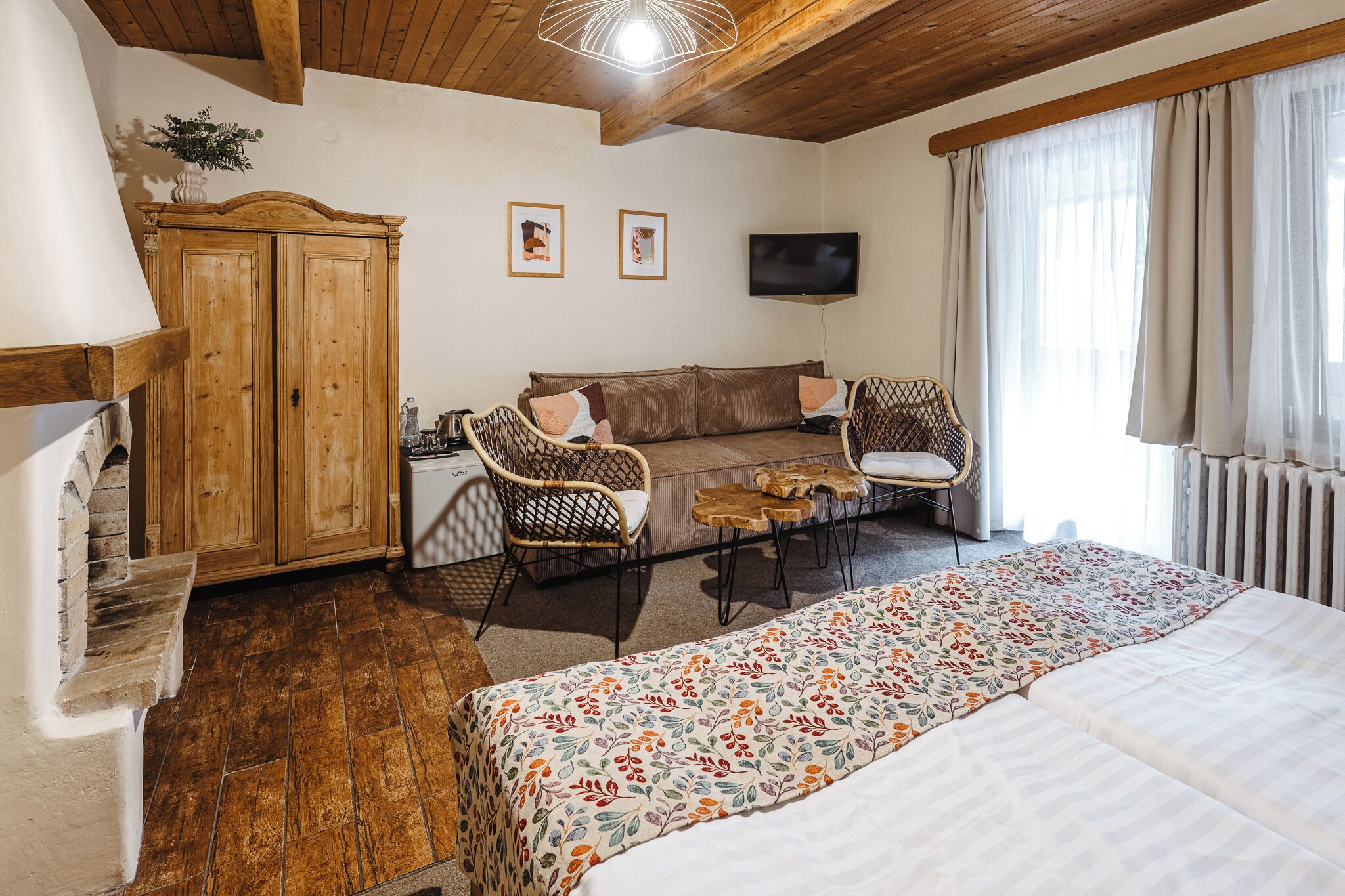 Una habitación acogedora con cama, sofá, dos sillones de mimbre, una mesita, un armario de madera y televisión en la pared.