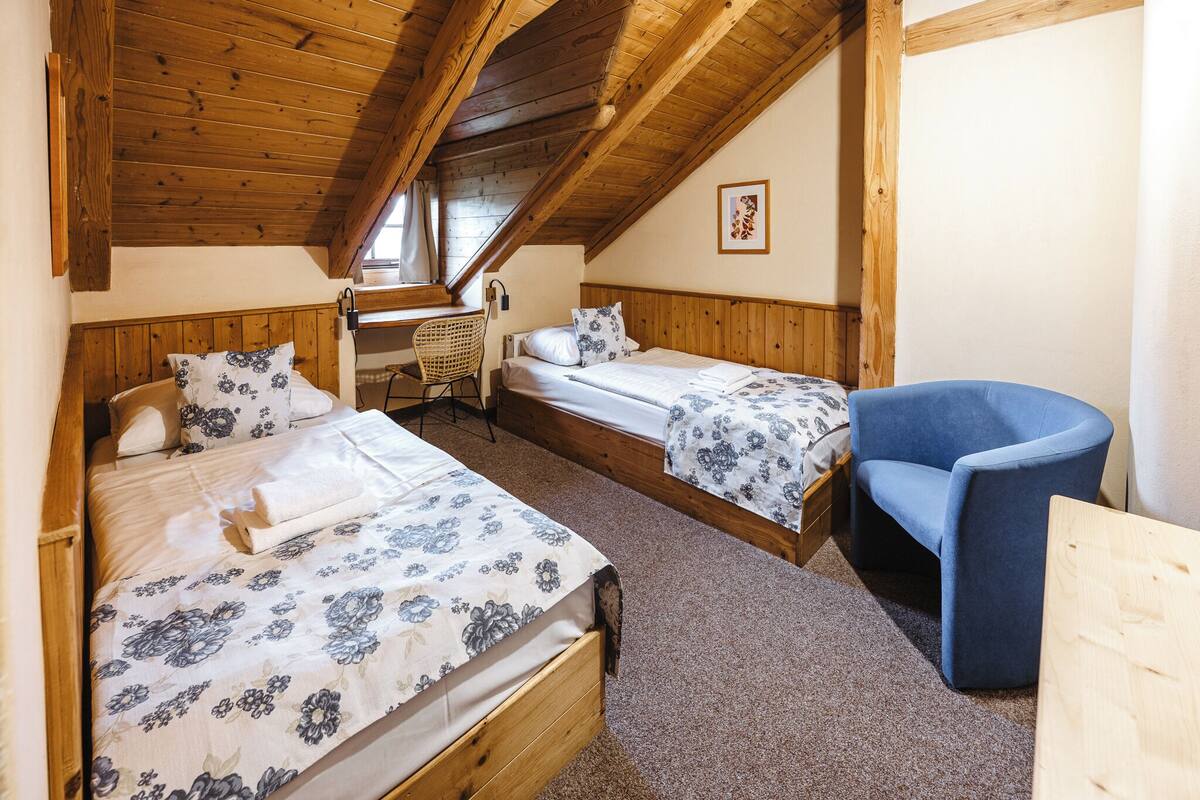A cozy attic bedroom with two beds, a blue armchair, and a wooden desk.