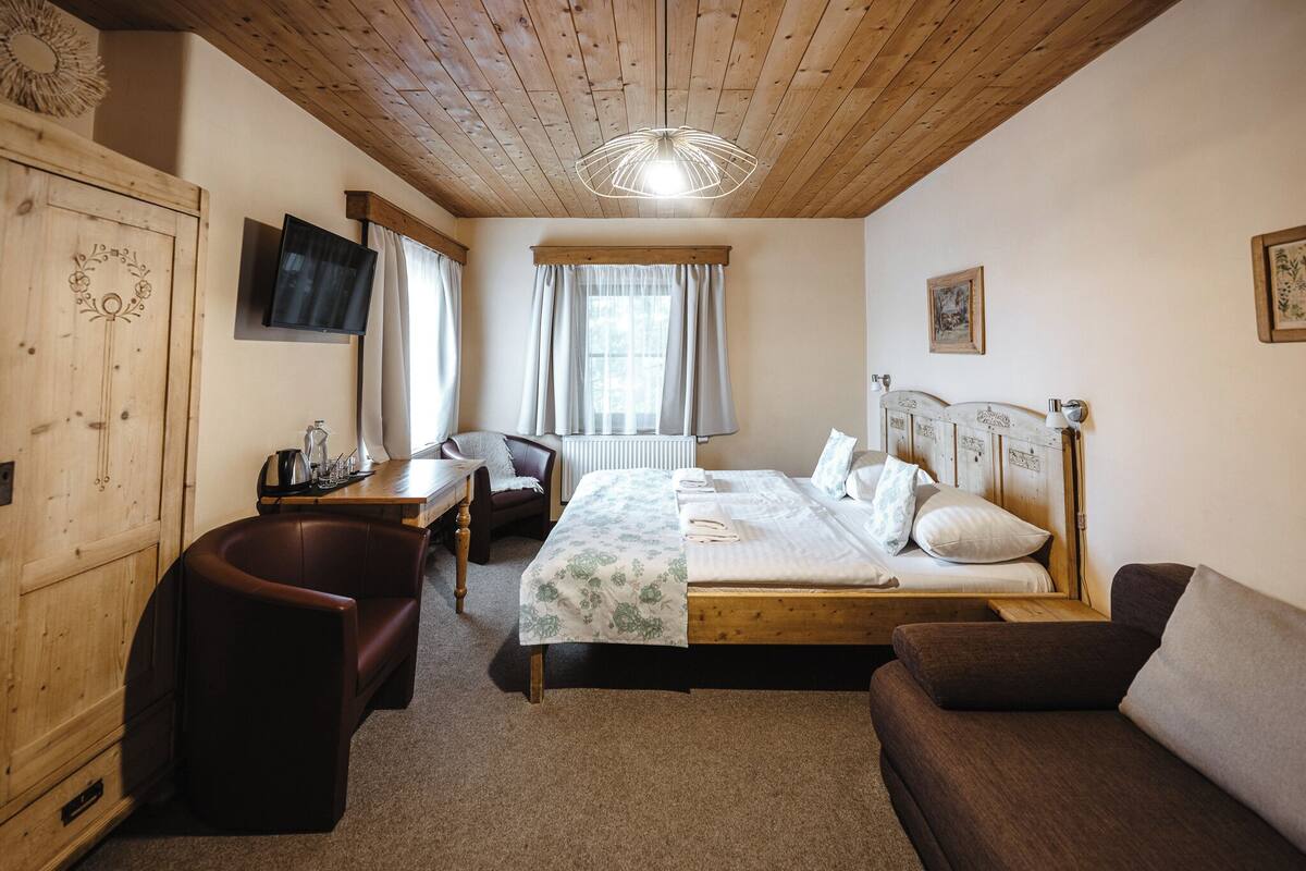 A cozy room with a wooden ceiling, a bed with a floral bedspread, a brown sofa, a desk with a chair, and a television on the wall.