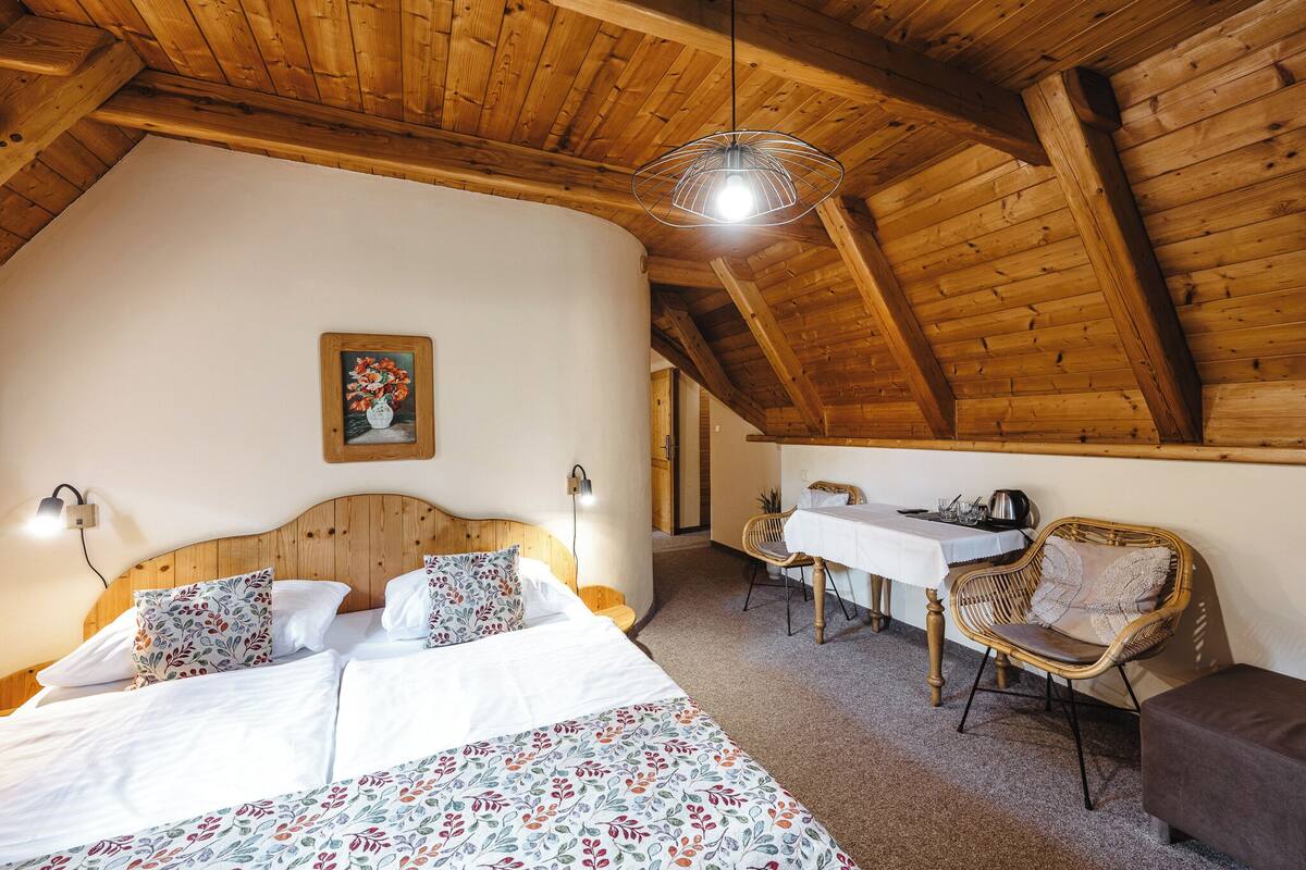 A cozy bedroom with a wooden ceiling, a bed with floral bedding, a dining table with chairs, and a painting on the wall.