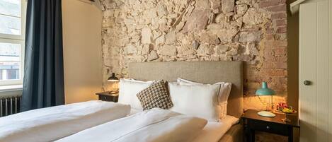 A double bed with white linens, a stone wall, a wooden ceiling beam, a nightstand with a lamp, and a small table with a fruit bowl.
