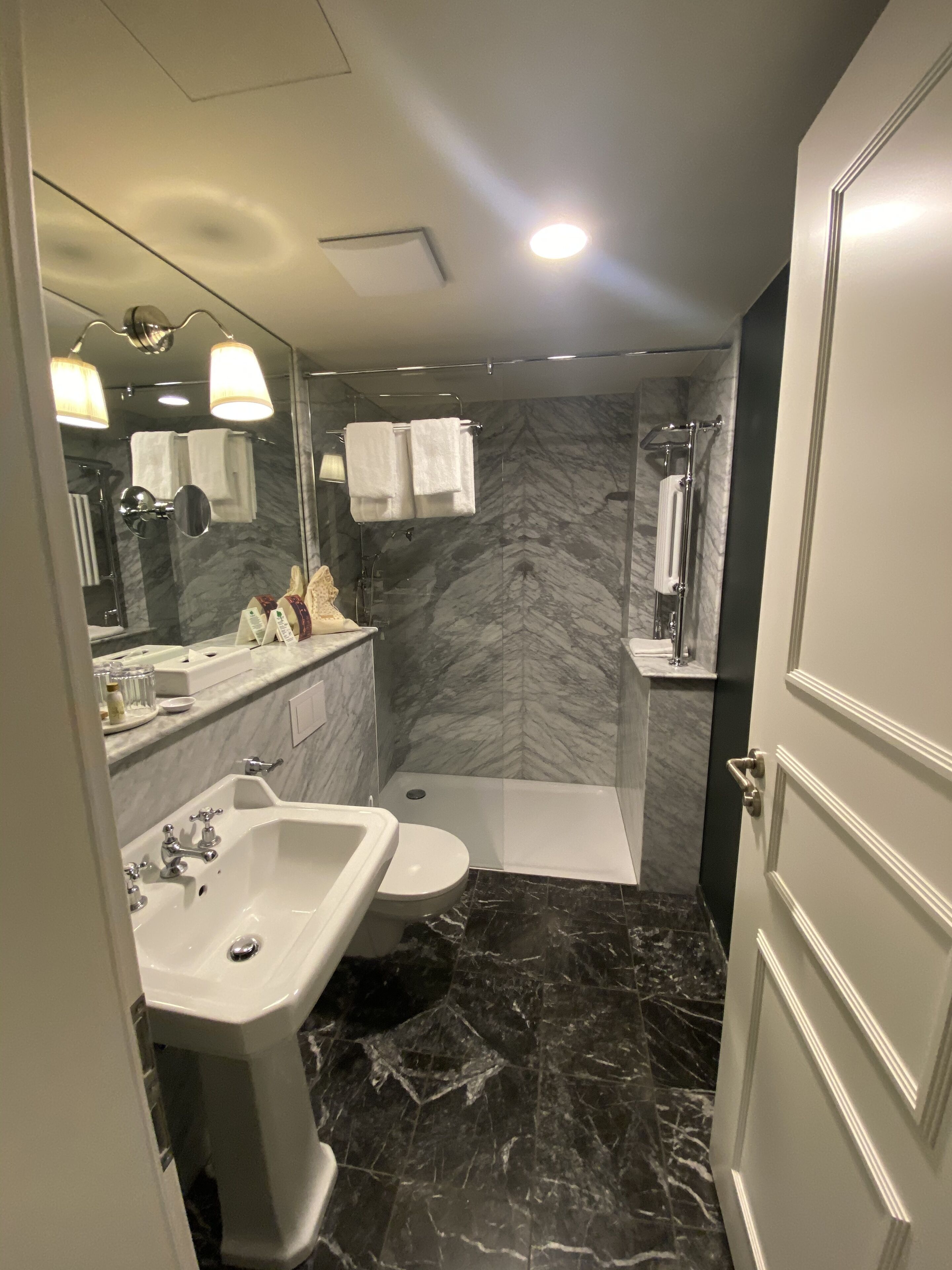 A modern bathroom with a marble floor, a sink, a toilet, a shower, and a mirror with lights.