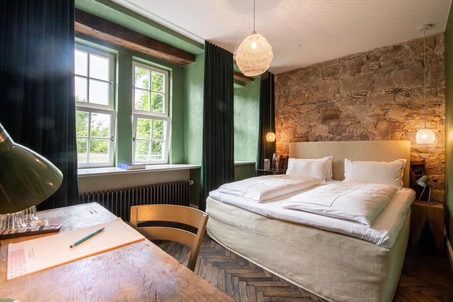A bedroom with a bed, a desk with a lamp, a chair, and a stone wall.