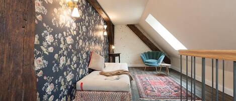 A loft bedroom with a sloped ceiling, a bed with a patterned bedspread, a blue chair, and a red and black rug.