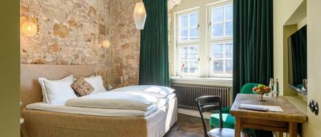 Un dormitorio con pared de piedra, una cama, un ventanal con cortinas, un radiador, una mesa de madera con una silla y un espejo.