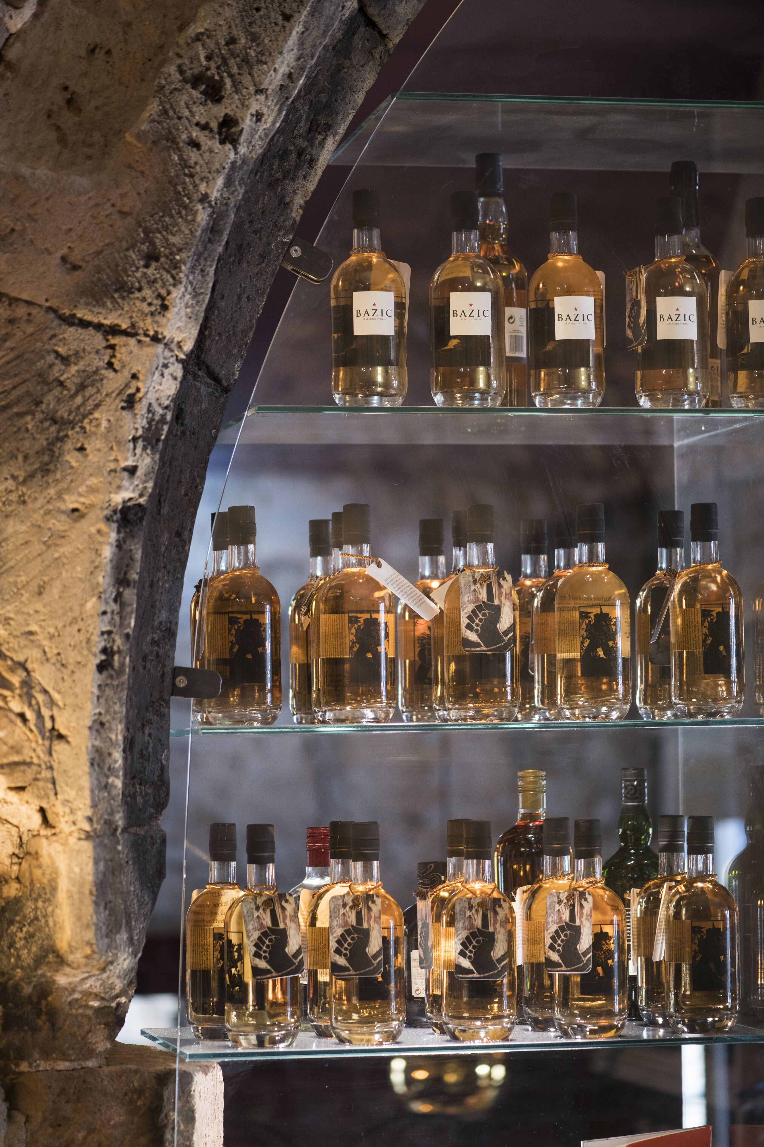 A display of various liquor bottles in a glass case with a stone wall background.