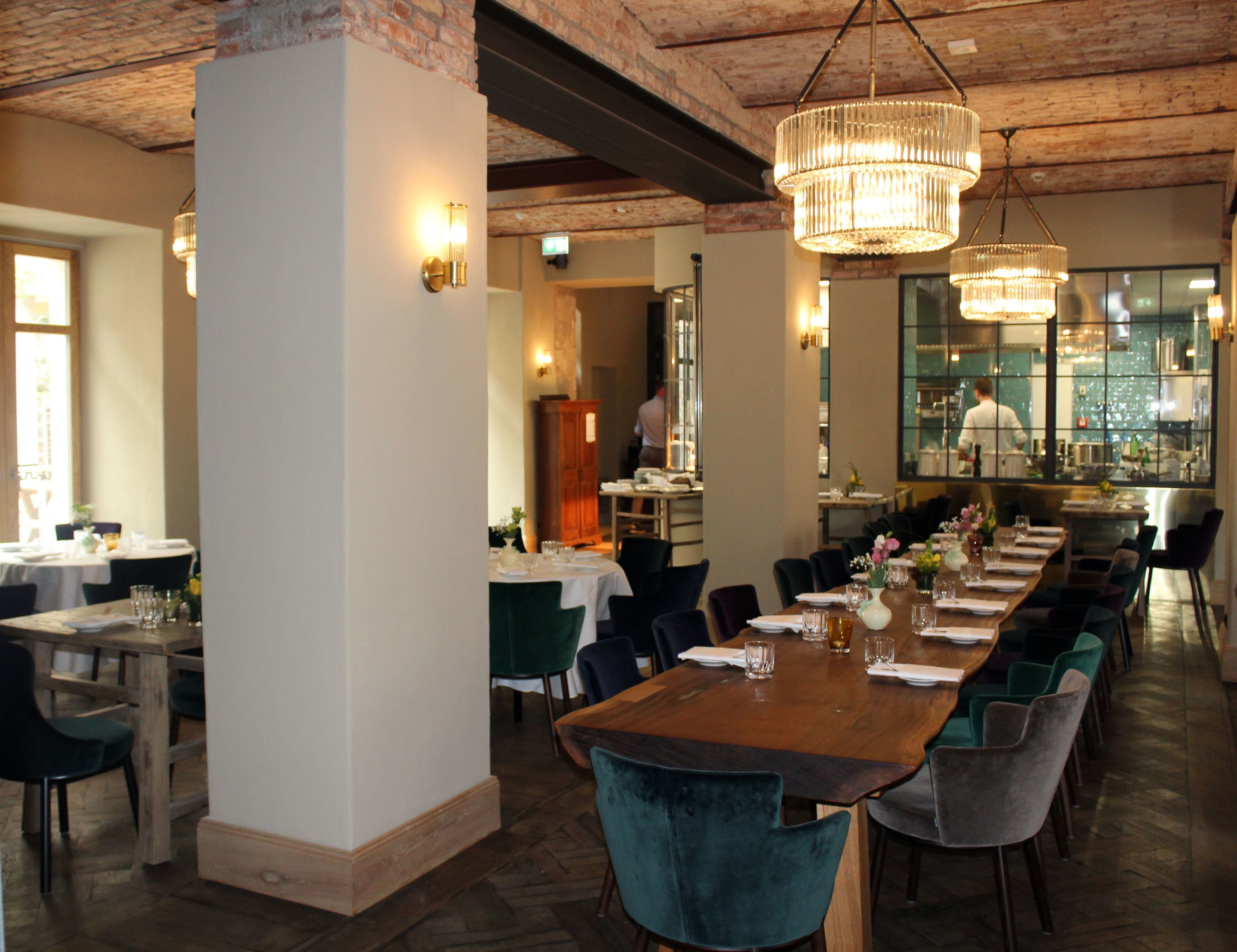 A restaurant with a long wooden table, green upholstered chairs, and hanging crystal chandeliers.