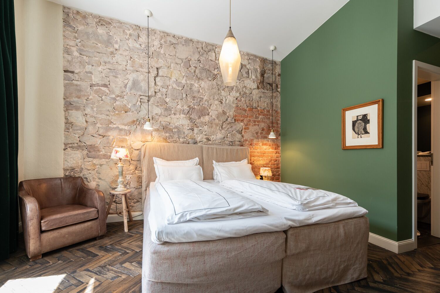A bedroom with a stone wall, a bed with white linens, a leather armchair, and a framed picture on the wall.