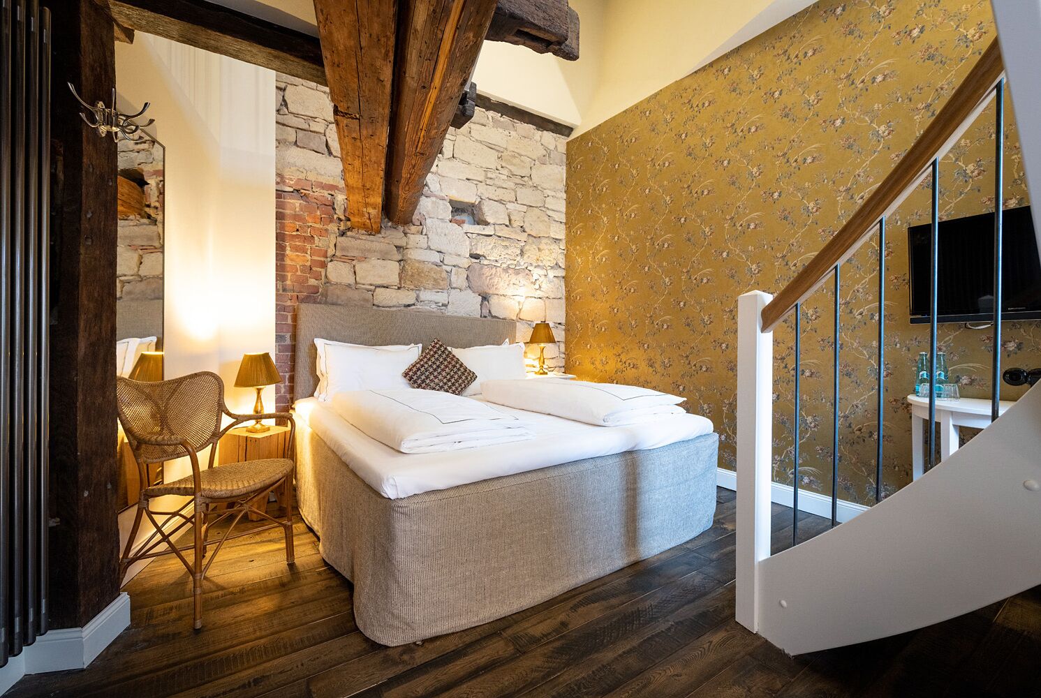 A bedroom with a stone wall, a bed with white linens, a wooden chair, and a lamp.