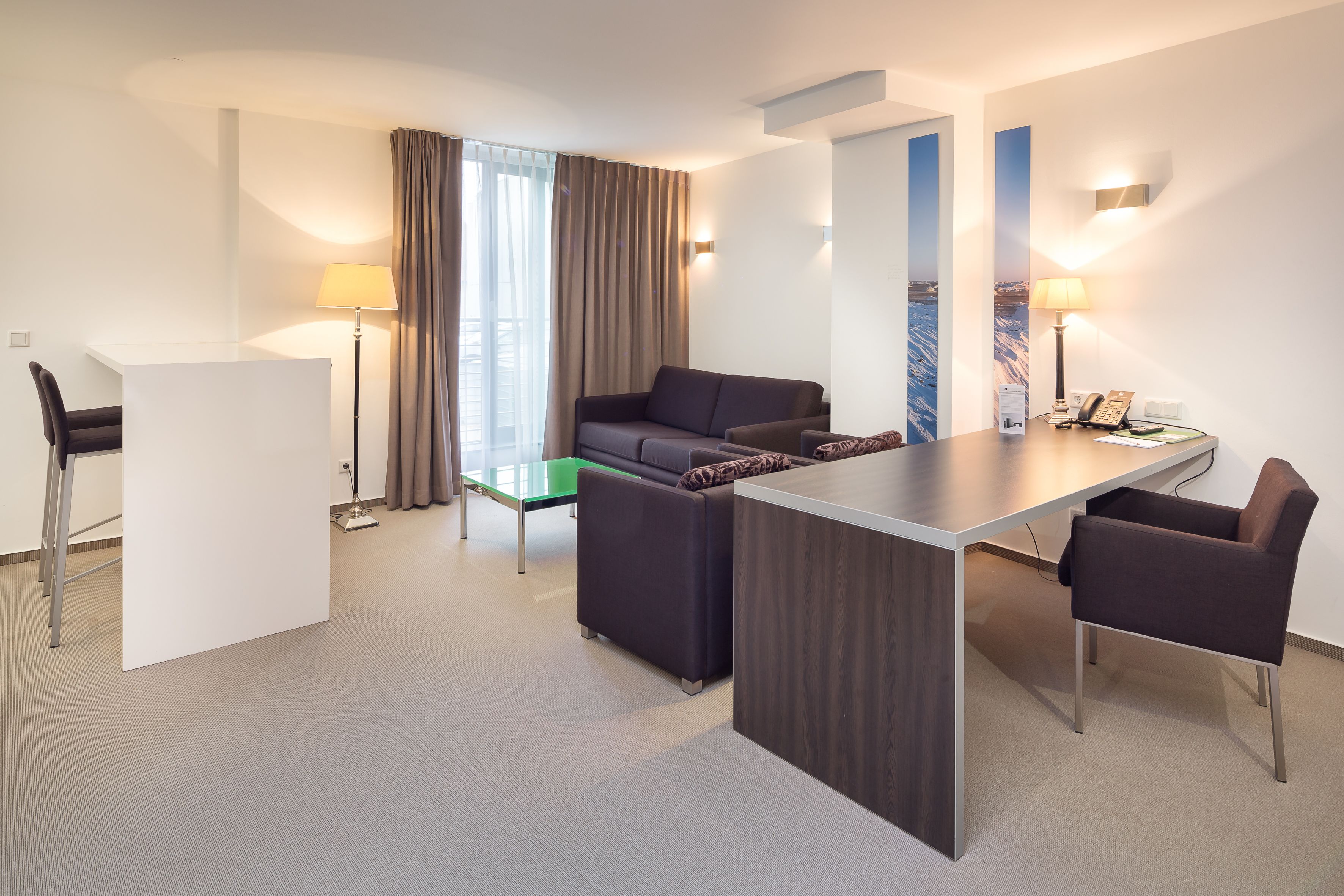 Een moderne hotelkamer met een bank, twee fauteuils, een bureau en een groene tafel.
