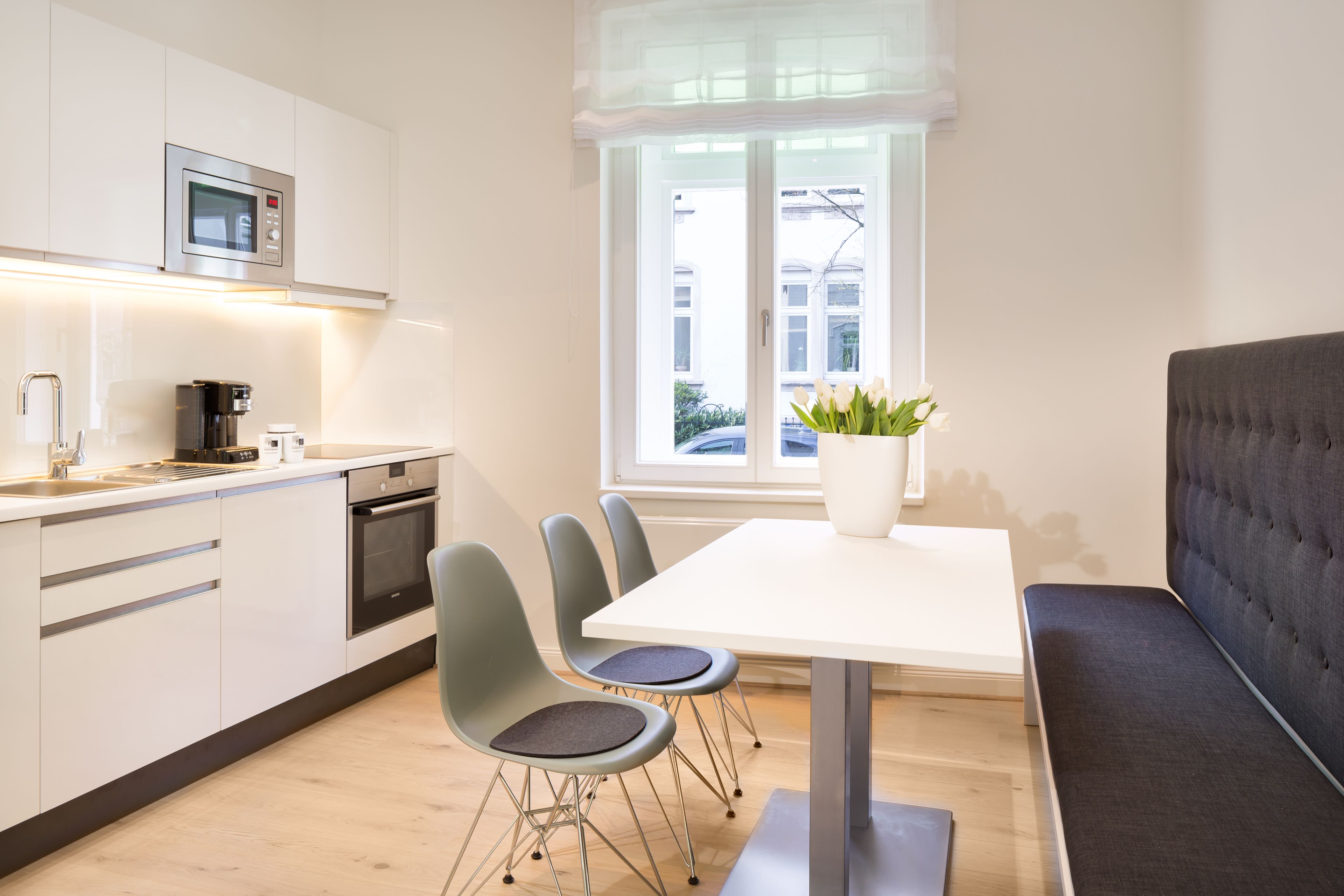 A modern kitchen with white cabinets, a microwave, and a coffee machine. There is a dining table with two chairs and a bench. A potted plant is on the table.