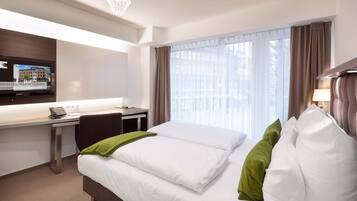 Una habitación de hotel con cama, escritorio, televisión y ventana con cortinas.