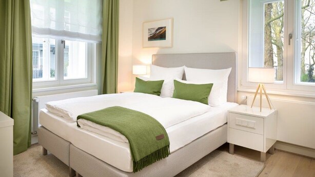 Een net opgemaakt bed met witte lakens en groene details, een nachtkastje met een lamp en een raam met gordijnen.