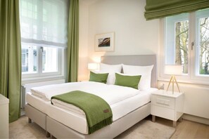 Una cama bien hecha con sábanas blancas y detalles verdes, una mesita de noche con una lámpara y una ventana con cortinas.