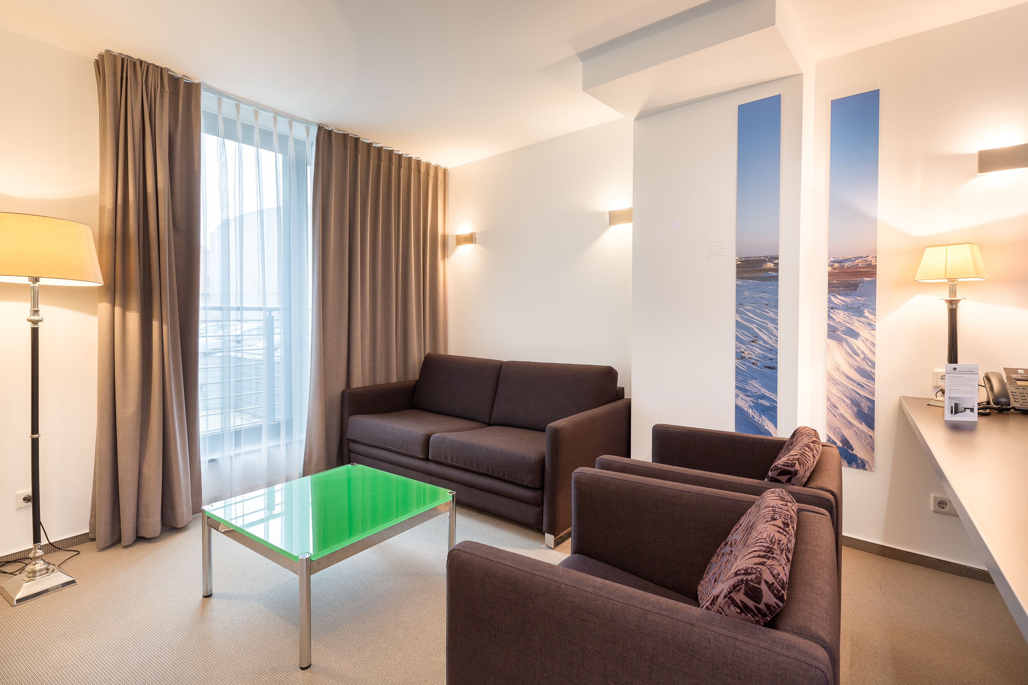 Una habitación de hotel moderna con sofá, sillones, una mesa de vidrio y un escritorio con lámpara.